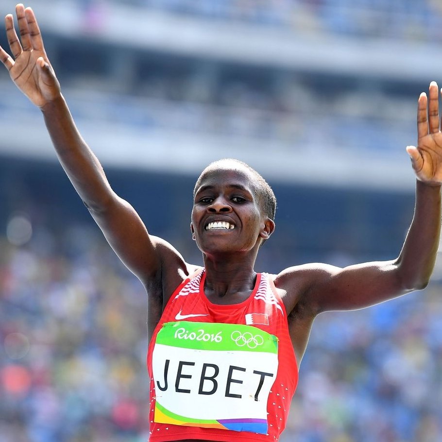 L'image montre une athlète célébrant sa victoire. Elle porte un débardeur rouge avec le nom "JEBET" inscrit dessus, et un logo des Jeux Olympiques sur sa tenue. Ses bras sont levés en signe de triomphe, et elle affiche un large sourire, exprimant la joie et l'excitation de son succès. En arrière-plan, on peut deviner une foule dans un stade, ajoutant à l'atmosphère festive de l'événement. L'éclairage est lumineux, mettant en valeur l'expression de bonheur sur son visage.
