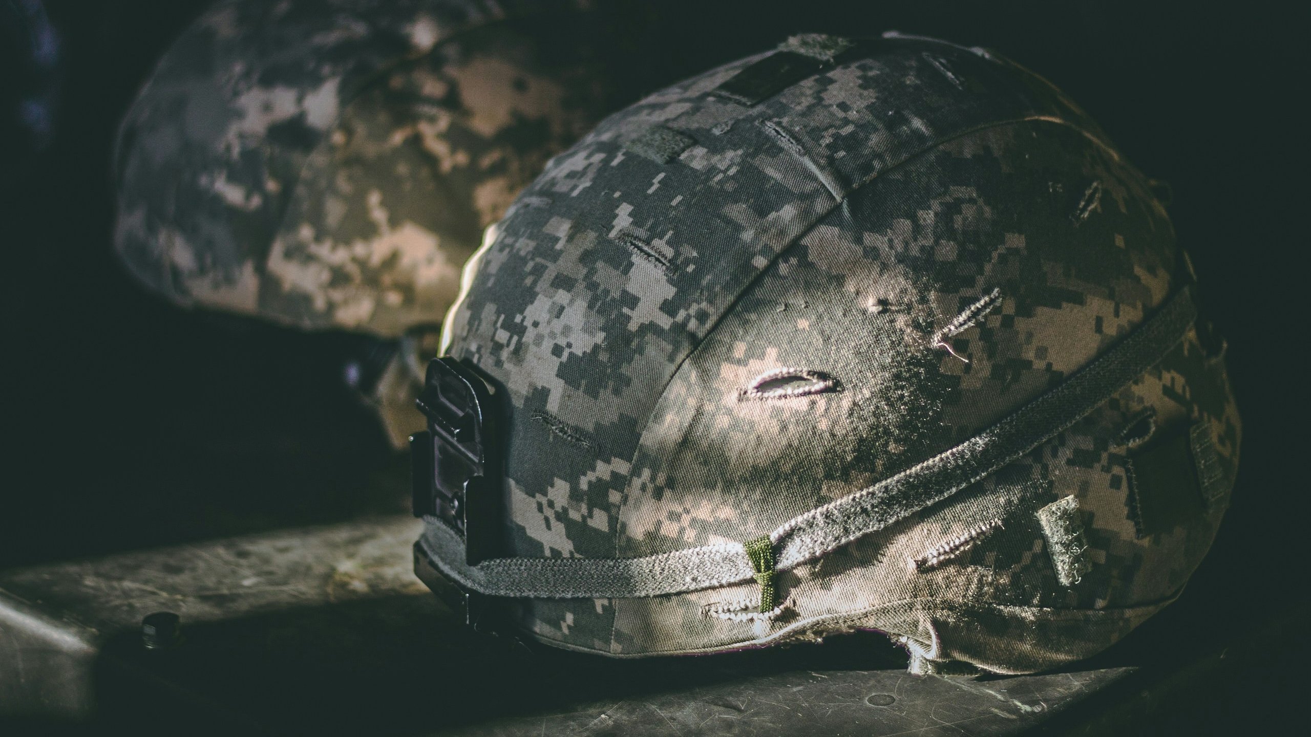 L'image montre deux casques militaires posés sur une surface sombre. Les casques sont de couleur camouflage, avec un motif de nuances grises et vertes. On peut voir des marques d'usure et des imperfections sur la surface, ce qui indique qu'ils ont été utilisés. La lumière frappe légèrement un des casques, mettant en évidence des textures et des détails, tandis que l'autre est dans l'ombre. L'ambiance générale est sobre et évoque un environnement militaire.