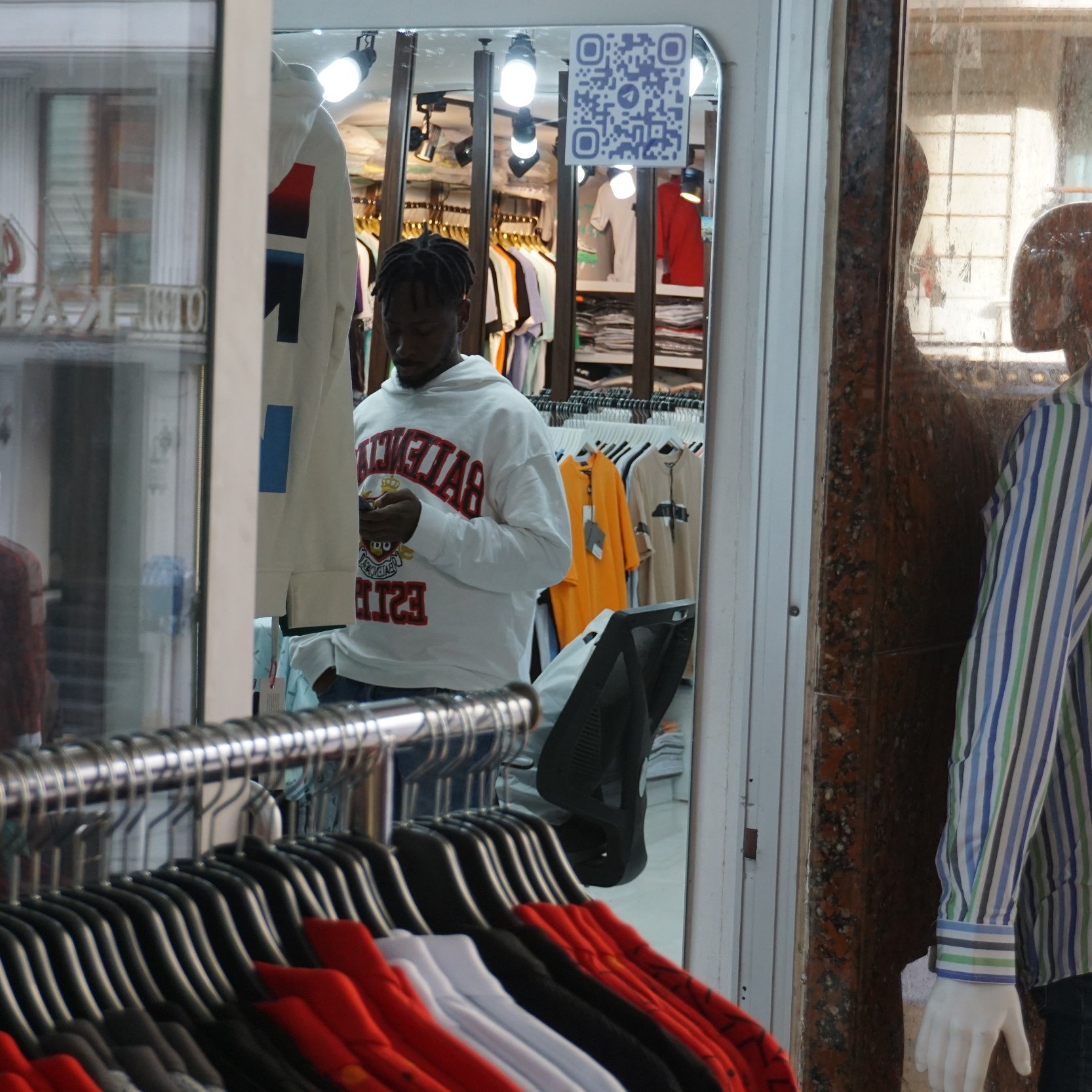 Dans cette image, nous voyons un magasin de vêtements. À l'avant, il y a un mannequin habillé d'une chemise rayée aux couleurs claires, affiché sur une vitrine. Derrière le mannequin, il y a un homme, qui porte un sweat à capuche blanc et semble utiliser son téléphone. Le magasin est bien éclairé, avec des étagères remplies de t-shirts colorés visibles à l'intérieur. Le fond de l'image montre une ambiance animée et des vêtements suspendus.