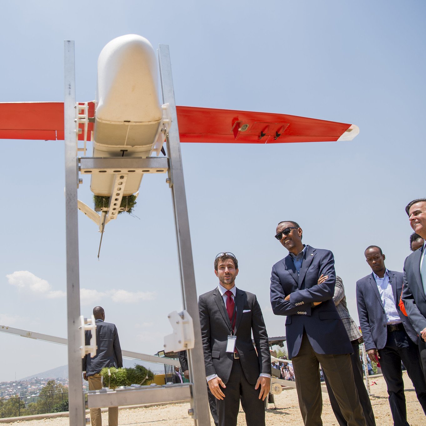 L'image montre un drone de forme allongée, aux ailes rouges et au corps blanc, placé sur une structure métallique. Plusieurs personnes, notamment des hommes en costumes, se tiennent à proximité, observant le drone avec intérêt. En arrière-plan, on aperçoit un paysage urbain et des collines sous un ciel bleu lumineux. L'atmosphère semble à la fois sérieuse et enthousiaste, suggérant un événement lié à la technologie ou à l'innovation.