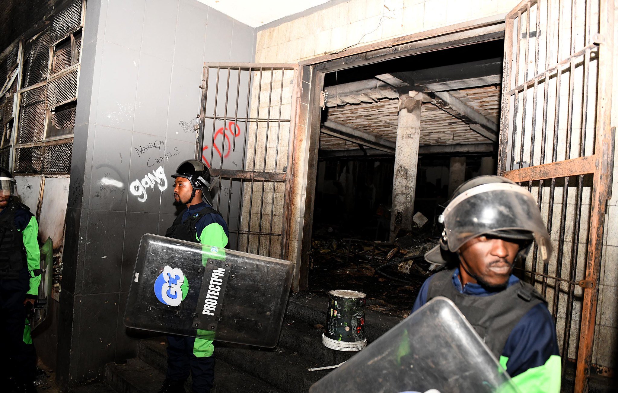 L'image montre une scène de sécurité, probablement après un événement perturbateur. On y voit deux agents de sécurité, vêtus d'un uniforme bleu et vert, portant des casques de protection et tenant des boucliers. Ils se tiennent devant une entrée, à côté de portes métalliques ouvertes. À l'intérieur, on aperçoit un espace sombre avec des traces de dégâts, comme des murs noircis. L'environnement semble chaotique, avec des débris visibles au sol. Des graffitis sont présents sur les murs, ajoutant une touche de désordre à la scène. L'atmosphère générale dégage un sentiment de tension et de vigilance.