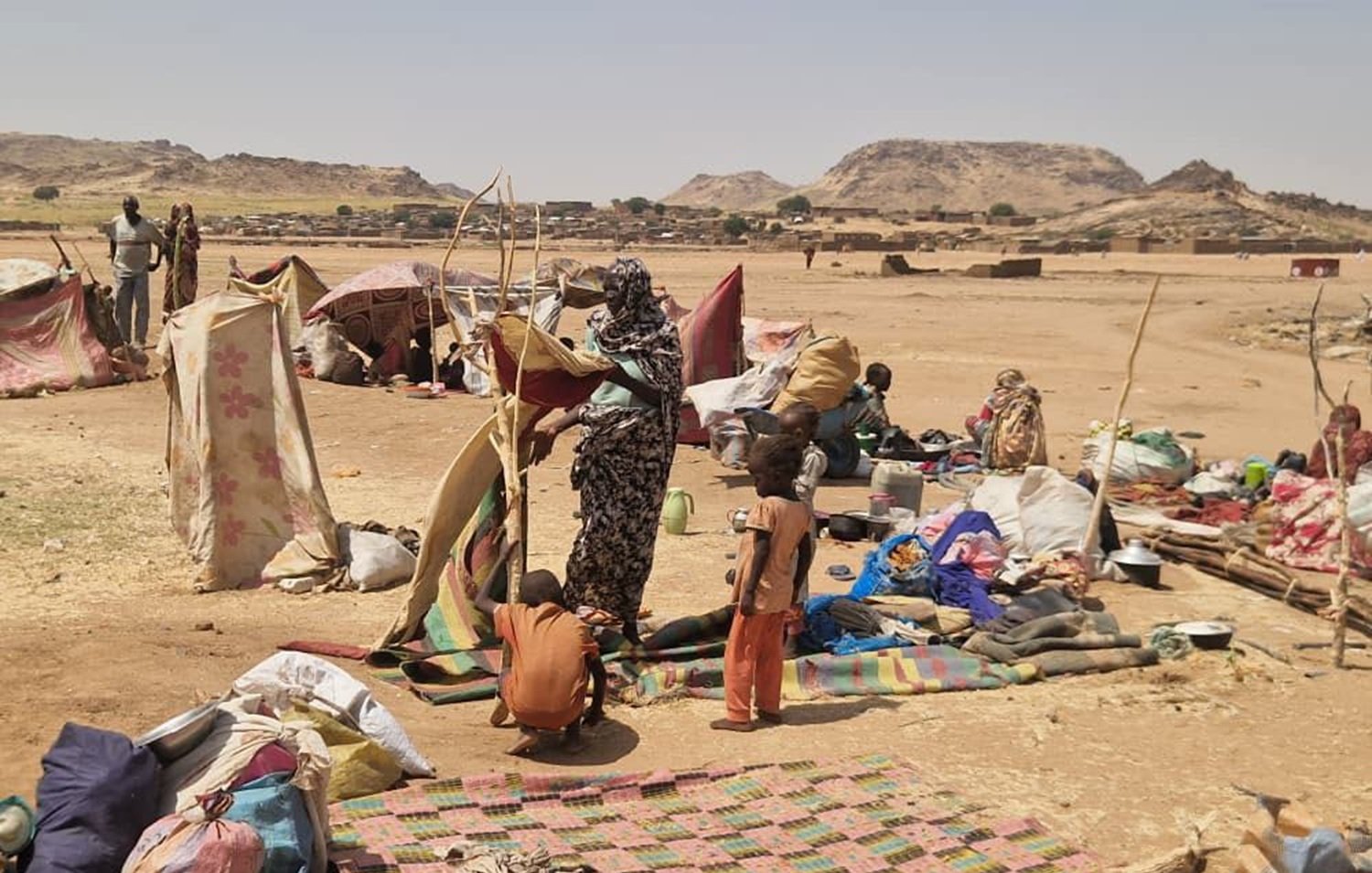 The image depicts a temporary settlement, likely a displaced persons camp or informal shelter in a rugged landscape. Several makeshift tents and tarps are set up, providing shelter for the residents. In the foreground, a woman is seen holding a structure, possibly a pole or support for one of the tents, while a child stands nearby. The ground is scattered with blankets, bags, and various belongings, suggesting a living area that is communal and somewhat crowded. The background shows a barren landscape with hills or rocky formations, indicating a harsh environment.