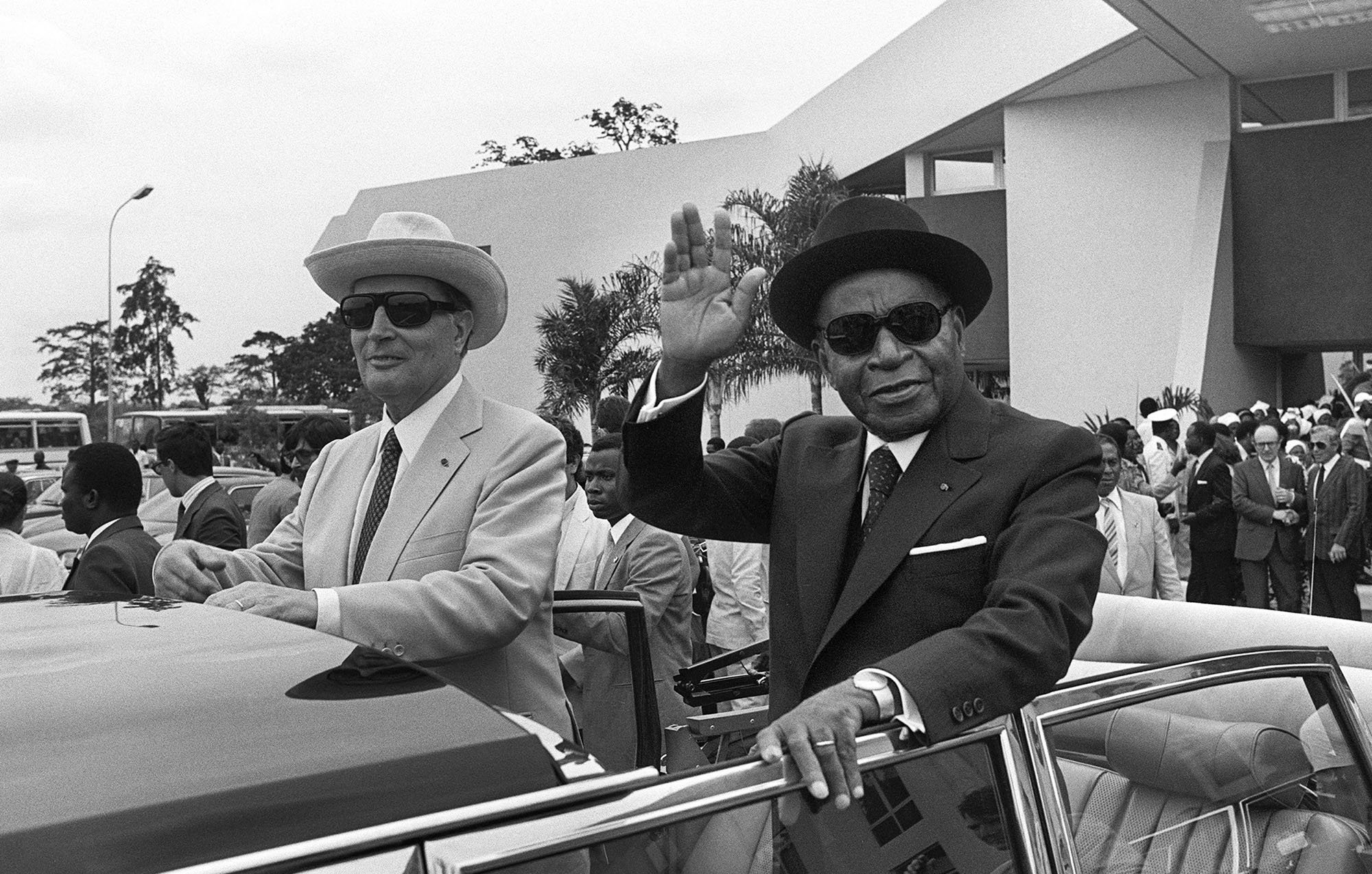 L'image représente deux hommes à l'extérieur d'une voiture décapotable. L'environnement semble être un événement officiel ou une cérémonie, avec des bâtiments modernes en arrière-plan. Le premier homme, à gauche, porte un costume clair, un chapeau de paille et des lunettes de soleil. Il a une expression sérieuse et regardent vers l'avant. Le second homme, à droite, est vêtu d'un costume foncé, d'un chapeau noir et également de lunettes de soleil. Il lève la main en signe de salut, avec un léger sourire. Il y a une foule de personnes en arrière-plan, habillées en costumes formels, ce qui indique une atmosphère festive ou cérémonielle. Des palmiers ou des arbres peuvent être aperçus, ajoutant une touche tropicale à l'environnement.