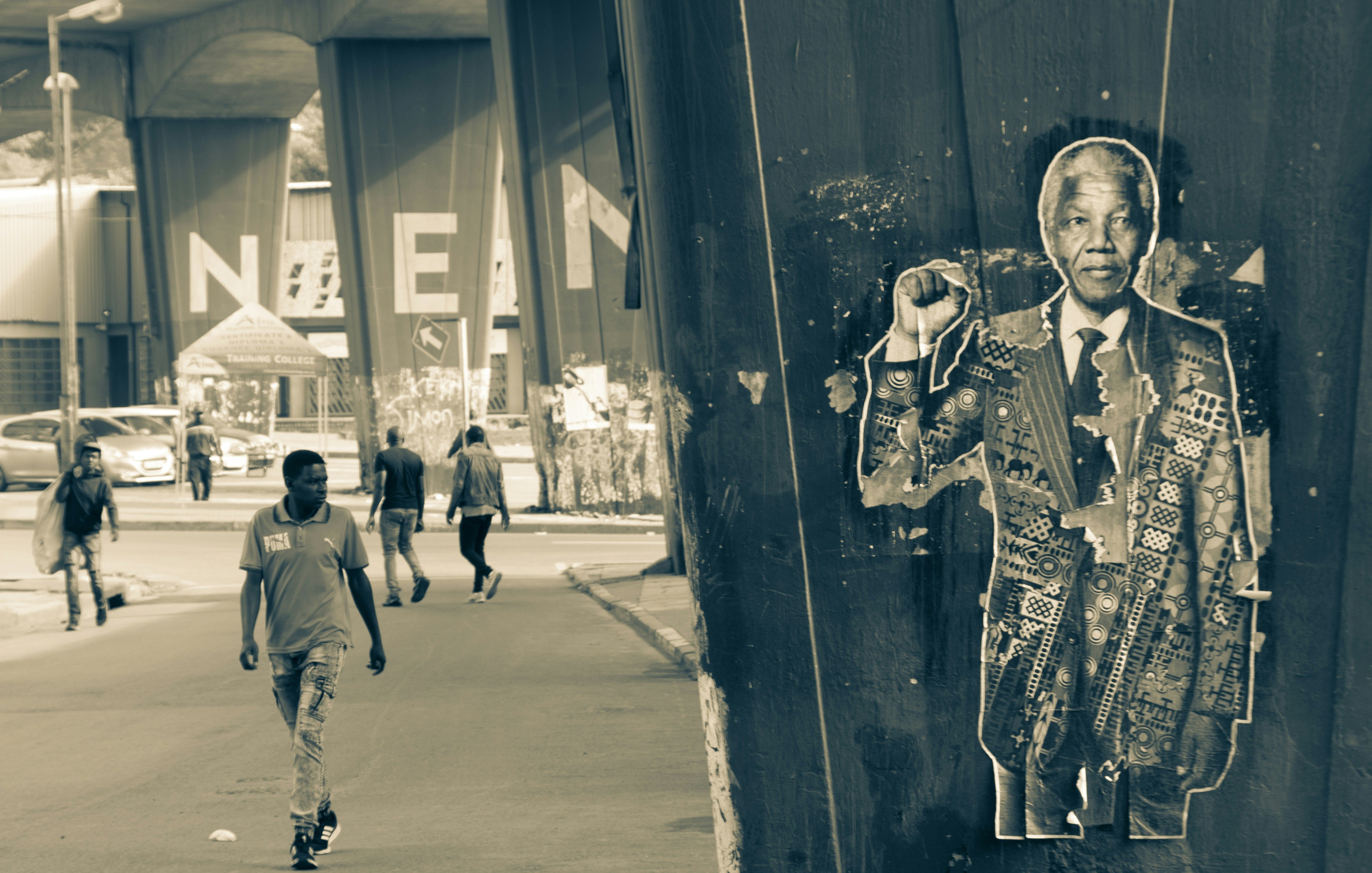 Dans cette image en noir et blanc, on voit une scène urbaine. À droite, un poster représentant Nelson Mandela est collé contre un grand pilier, le montrant avec un poing levé en signe de résistance. Le poster est texturé et présente des motifs. À gauche, un homme marche sur un chemin urbain, entouré d'autres personnes, qui semblent se diriger vers un lieu dont on aperçoit le nom inscrit en grandes lettres « NEAM » à l'arrière-plan. L'atmosphère de la scène évoque un mélange d'histoire, de mémoire et de vie quotidienne dans un environnement animé.