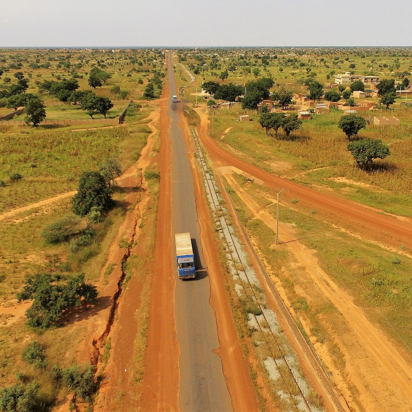 Cette image présente un paysage rural étendu, où une route goudronnée traverse une vaste plaine. Sur la droite, on aperçoit un camion bleu qui circule, indiquant une activité de transport. De part et d'autre de la route, le sol est d'un rouge terreux, typique de certaines régions africaines, semé de petites collines et de végétation éparse, notamment des arbres et des buissons. Sur la gauche de l'image, on peut distinguer des habitations simples, suggérant la présence d'un village. L'arrière-plan est composé de champs et d'arbres, renforçant l'impression de tranquillité et d'espace ouvert. L'ensemble évoque une atmosphère de ruralité, avec une nature généreuse, baignée par une lumière intense.