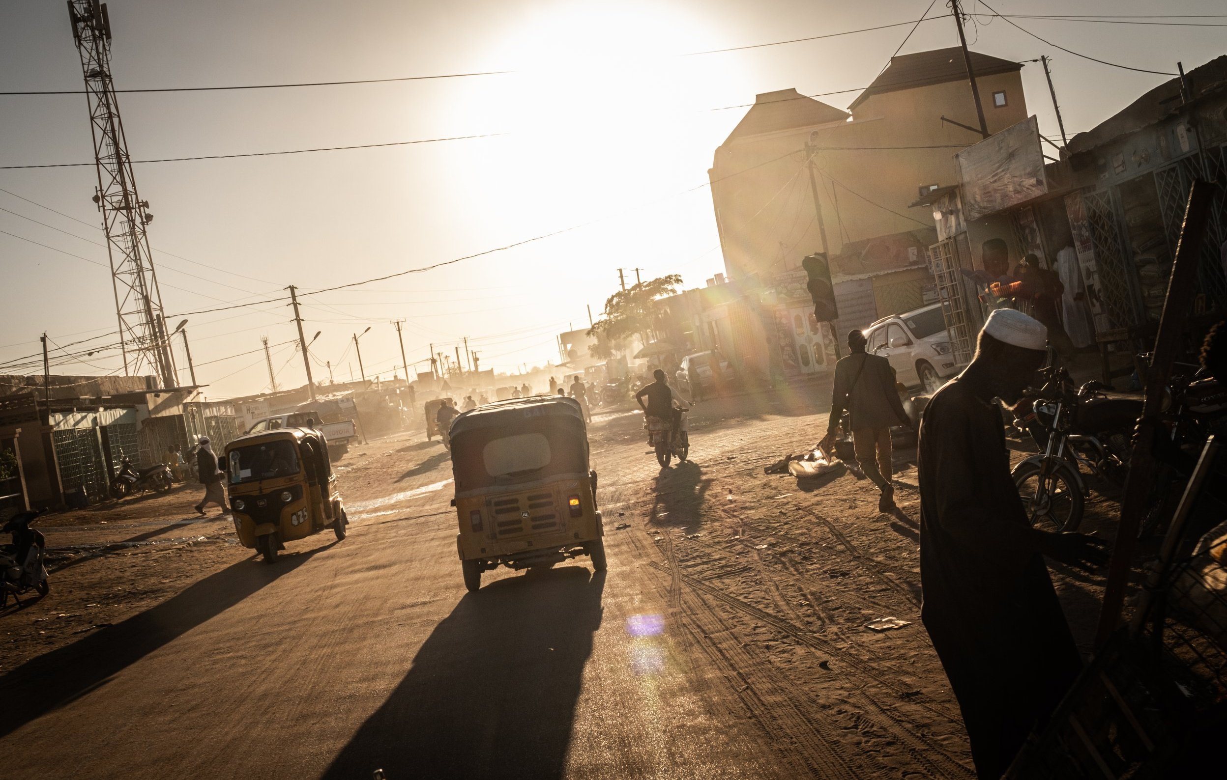 The image depicts a quiet, sandy street at sunset, where the golden light of the sun is visible on the horizon. On one side, there are simple, earth-toned walls of buildings, casting long shadows in the warm light. A few individuals are scattered along the street, with one person sitting and another standing near a donkey. The atmosphere suggests a peaceful, rural environment, with minimal traffic and a sense of tranquility as day transitions to night.