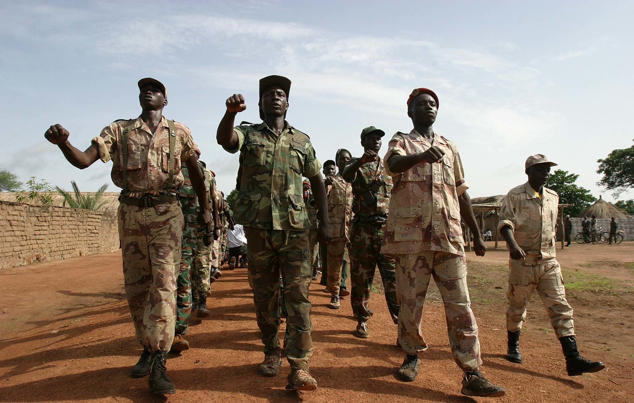 L'image montre un groupe de soldats marchant en formation sur un sol poussiéreux. Ils portent des uniformes militaires variés, allant du camouflé au beige, qui suggèrent une ambiance militaire. Certains soldats brandissent le poing en signe de détermination, tandis que d'autres avancent d'un pas ferme. En arrière-plan, on peut voir des tentes militaires ou des structures rudimentaires, suggérant un camp. Le ciel est légèrement nuageux, laissant passer quelques rayons de soleil. L'atmosphère générale évoque la discipline et la camaraderie au sein de ce groupe.