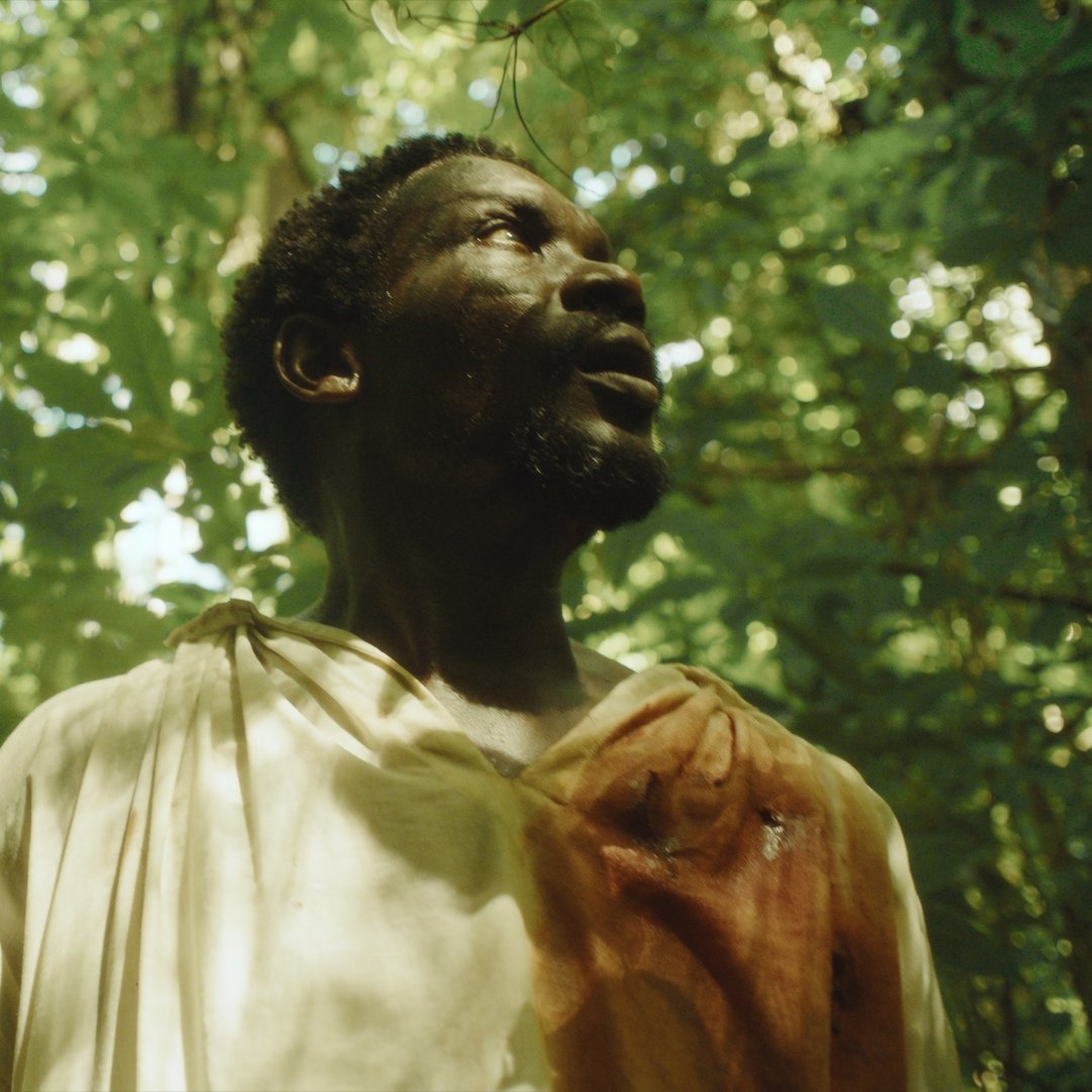 L'image montre un homme debout dans une jungle luxuriante. Il porte une tunique légère, dont une partie est tachée de rouge, suggérant un événement émotionnel ou tragique. Le visage de l'homme exprime la surprise ou l'inquiétude, avec des traits marqués par la lumière douce qui filtre à travers le feuillage dense. Les feuilles vertes et les ombres créent une atmosphère à la fois mystérieuse et intense, plongeant le spectateur dans l'ambiance d'une nature vibrante et sauvage. On peut imaginer les sons de la jungle, comme le chant des oiseaux et le bruissement des feuilles, qui entourent l'homme.