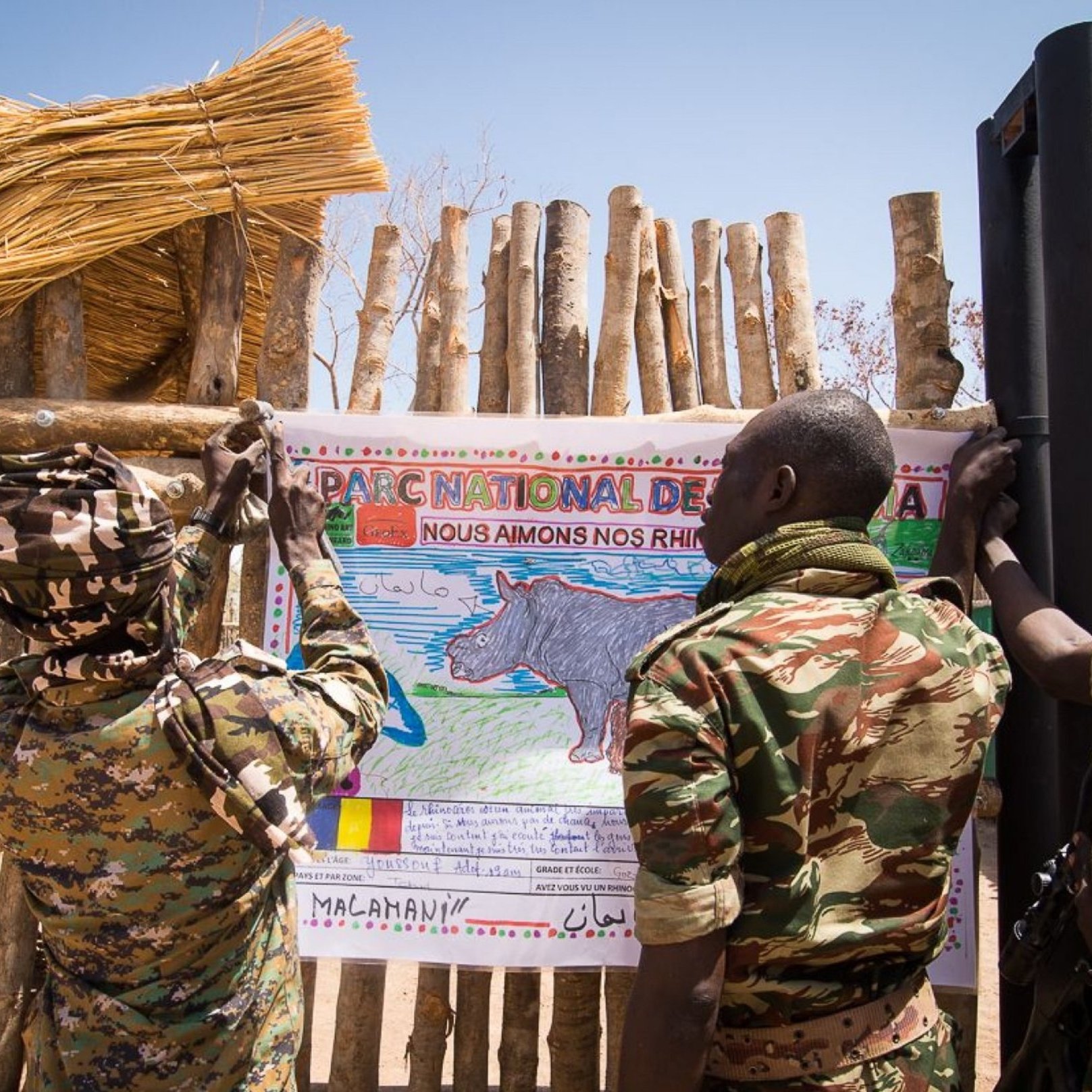 Sur l'image, nous voyons deux soldats en uniforme coloré, engagés dans une action collective. Ils sont en train d'accrocher une grande affiche sur un mur en bois fait de branches. L'affiche représente un rhinocéros, avec un dessin coloré et un message écrit en français qui exprime un engagement envers la protection de cette espèce. Le cadre est en plein air, avec un ciel bleu et quelques éléments naturels visibles. L'atmosphère dégage un sentiment de protection et d'éducation environnementale, soulignant l'importance de préserver la faune.