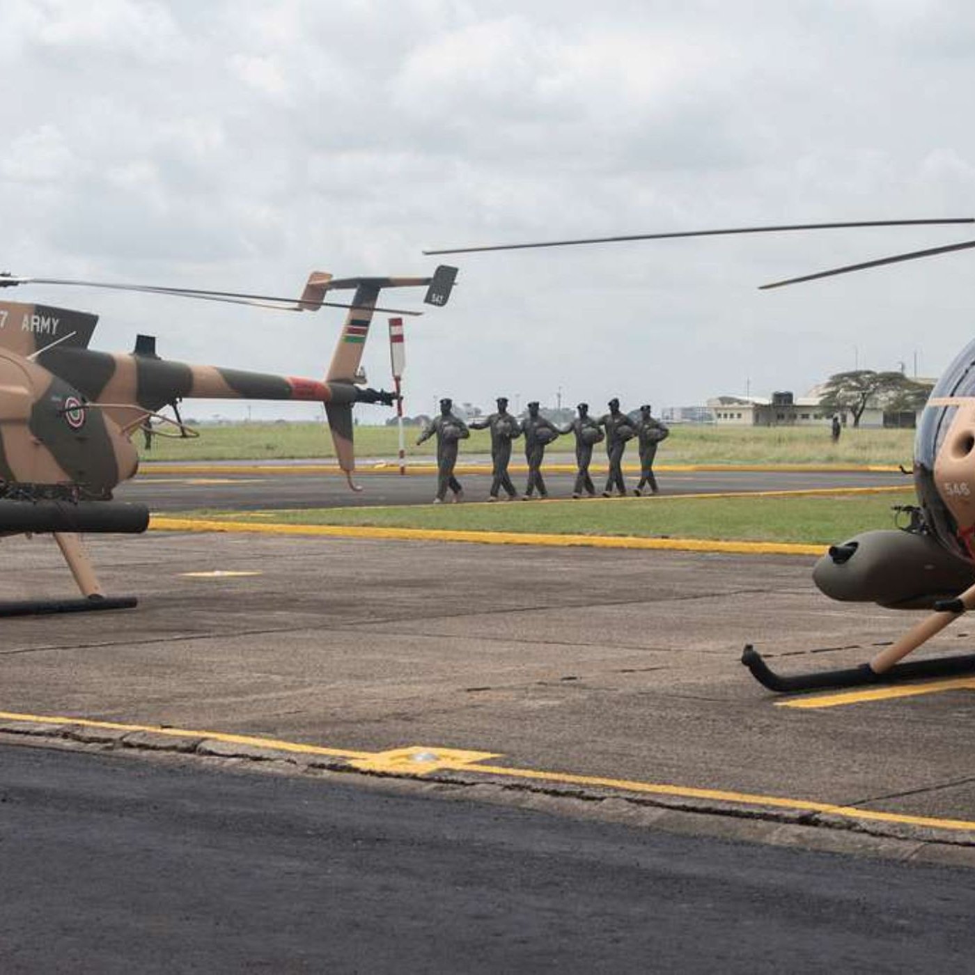 L'image montre une scène aérienne sur un terrain d'aviation. Deux hélicoptères, de couleur beige avec des motifs camouflage, sont stationnés sur le tarmac. À l'arrière-plan, un groupe de soldats en uniforme se dirige en formation vers un point de destination, créant une atmosphère dynamique et militaire. Le ciel est nuageux, ajoutant une ambiance dramatique à la scène. Le paysage autour est dégagé, avec des bâtiments épars visibles au loin.
