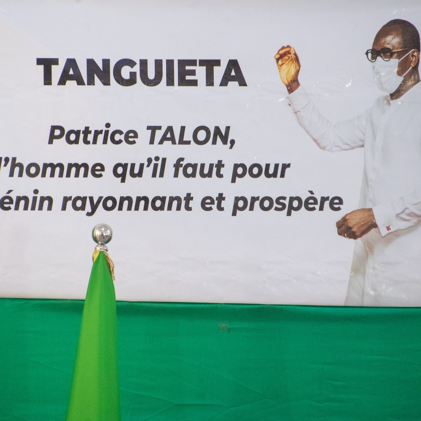 L'image présente une affiche accrochée sur un fond vert. Cette affiche met en avant le nom "TANGUIETA" en haut, suivi d'une mention concernant Patrice TALON. Le texte indique qu'il est "l'homme qu'il faut pour un Bénin rayonnant et prospère." On peut également voir un homme en tenue blanche, portant un masque. Il est positionné à droite de l'affiche, levant une main dans un geste de discours ou de présentation. À gauche de l'affiche, se trouve un drapeau vert qui se dresse à la verticale. L'ensemble véhicule un message d'espoir et de prospérité pour le pays.