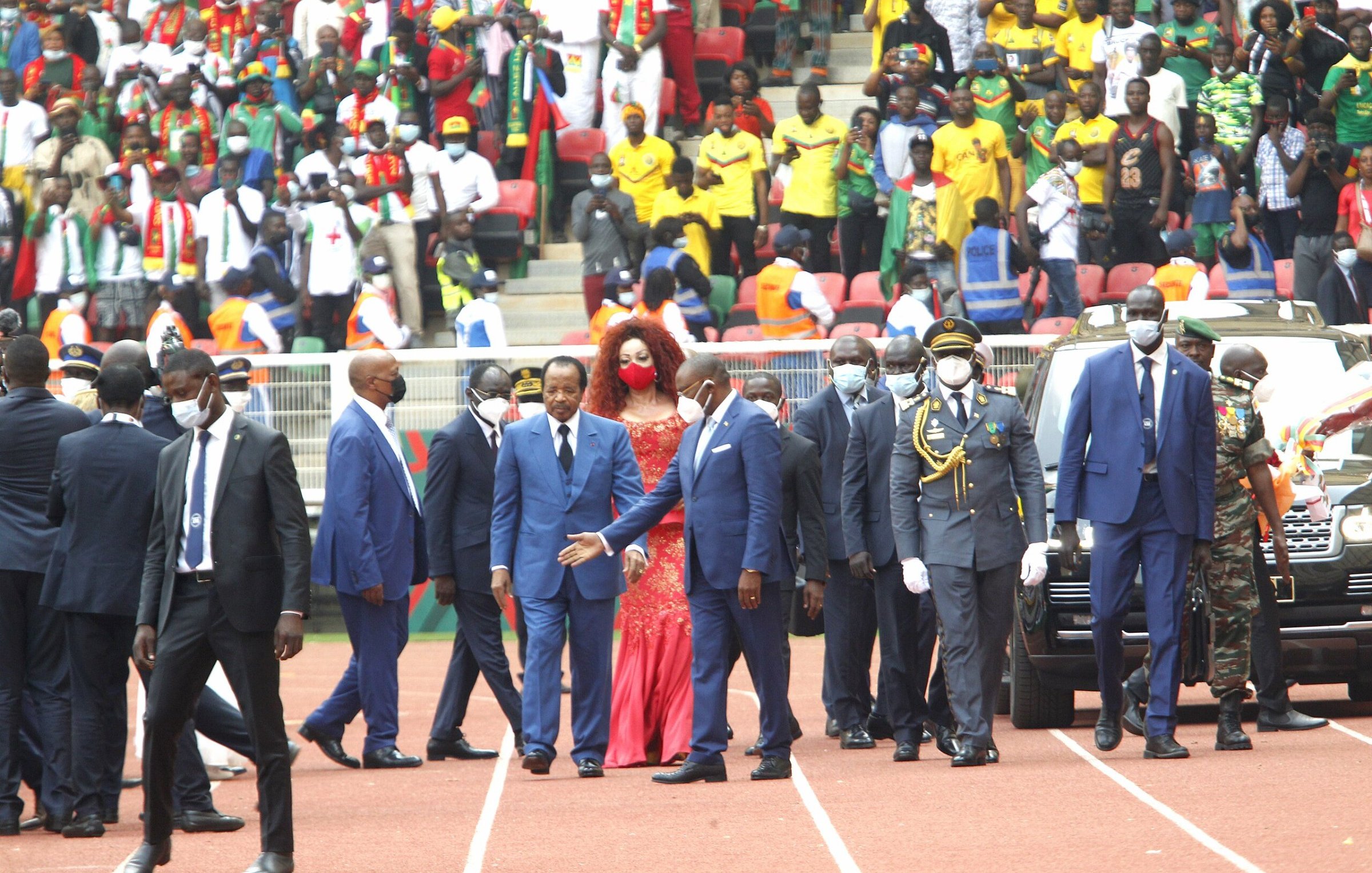 L'image montre un événement en extérieur, probablement un stade rempli de spectateurs. Au premier plan, un groupe de personnalités, dont plusieurs hommes en costume sombre et cravate, marche avec assurance. Parmi eux, une femme se distingue grâce à sa robe rouge éclatante et sa coiffure volumineuse. Ils semblent être en route pour un moment important, entourés de sécurité. À l'arrière-plan, on peut deviner une foule colorée et animée, avec des supporters portant divers vêtements aux couleurs vives, témoignant d'une atmosphère festive. L'ambiance générale semble être celle d'une grande célébration ou d'une cérémonie officielle.