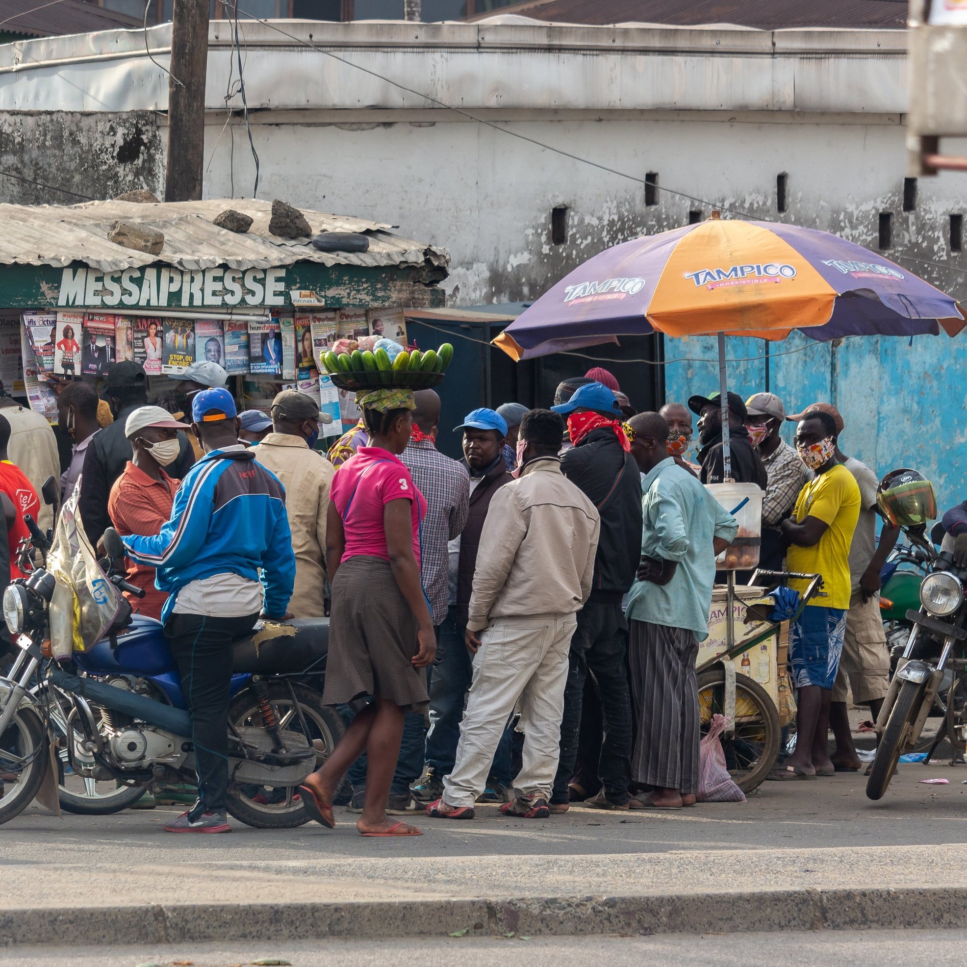 L'image montre une scène animée dans une rue d'une ville. On peut voir un groupe de personnes rassemblées autour d'un kiosque à journaux, qui semble être un lieu de rencontre. Certaines personnes portent des masques, ce qui indique une attention à la santé publique. À l'avant, une femme porte un grand panier de bananes sur sa tête, ce qui met en valeur une pratique courante dans de nombreuses cultures. Des motos et une voiture jaune sont garées à proximité, ajoutant à l'ambiance urbaine. En arrière-plan, on peut distinguer des bâtiments simples, avec des couleurs plutôt ternes, ce qui donne un aspect de vie quotidienne dans ce quartier.