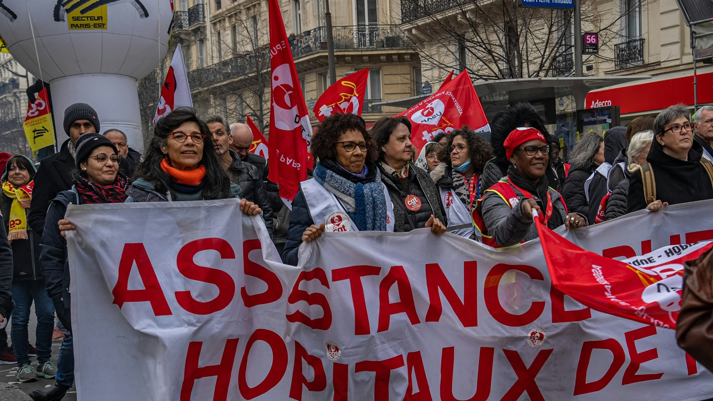 L'image montre une manifestation dans une rue animée. Au premier plan, un groupe de personnes tient une grande banderole blanche sur laquelle est écrit "ASSISTANCE HÔPITAUX DE…" en lettres rouges. Les manifestants arborent des sourires et semblent déterminés. On aperçoit également plusieurs drapeaux rouges, qui sont souvent associés à des syndicats. L'atmosphère est dynamique, avec une multitude de personnes autour, représentant la solidarité et l'engagement. Les bâtiments en arrière-plan suggèrent une ambiance urbaine, typique des grandes villes.