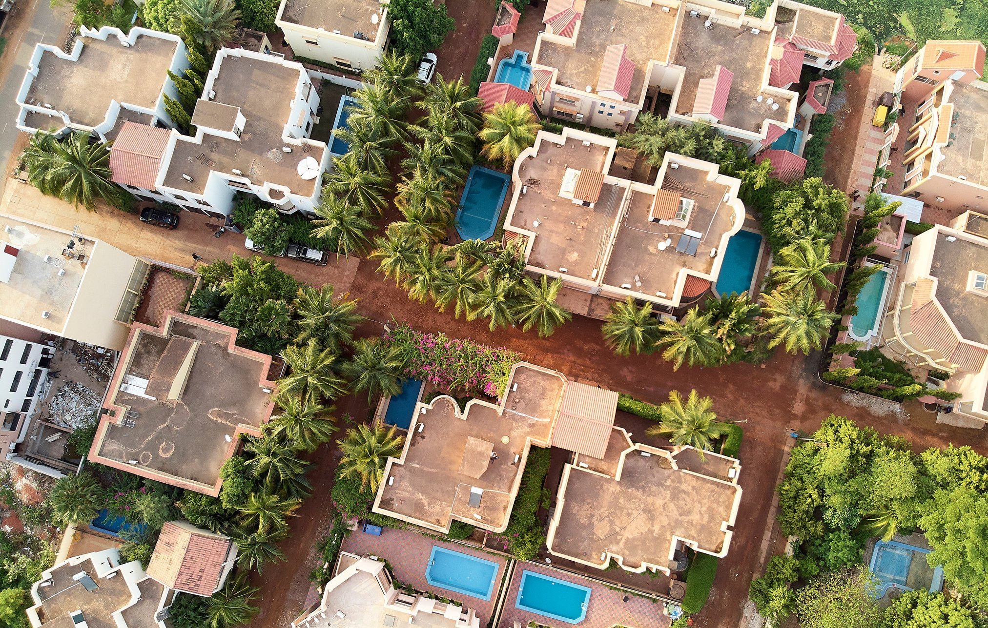 L'image montre une vue aérienne d'un complexe résidentiel. On y voit plusieurs maisons aux toits plats, disposées autour de petites piscines bleues. Parmi ces maisons, il y a de nombreuses palmiers qui ajoutent une touche de verdure. Les allées sont en terre et les maisons sont agencées de manière à créer un réseau urbain harmonieux. Les couleurs de la terre et des toits se marient pour donner une ambiance chaleureuse et accueillante au paysage.