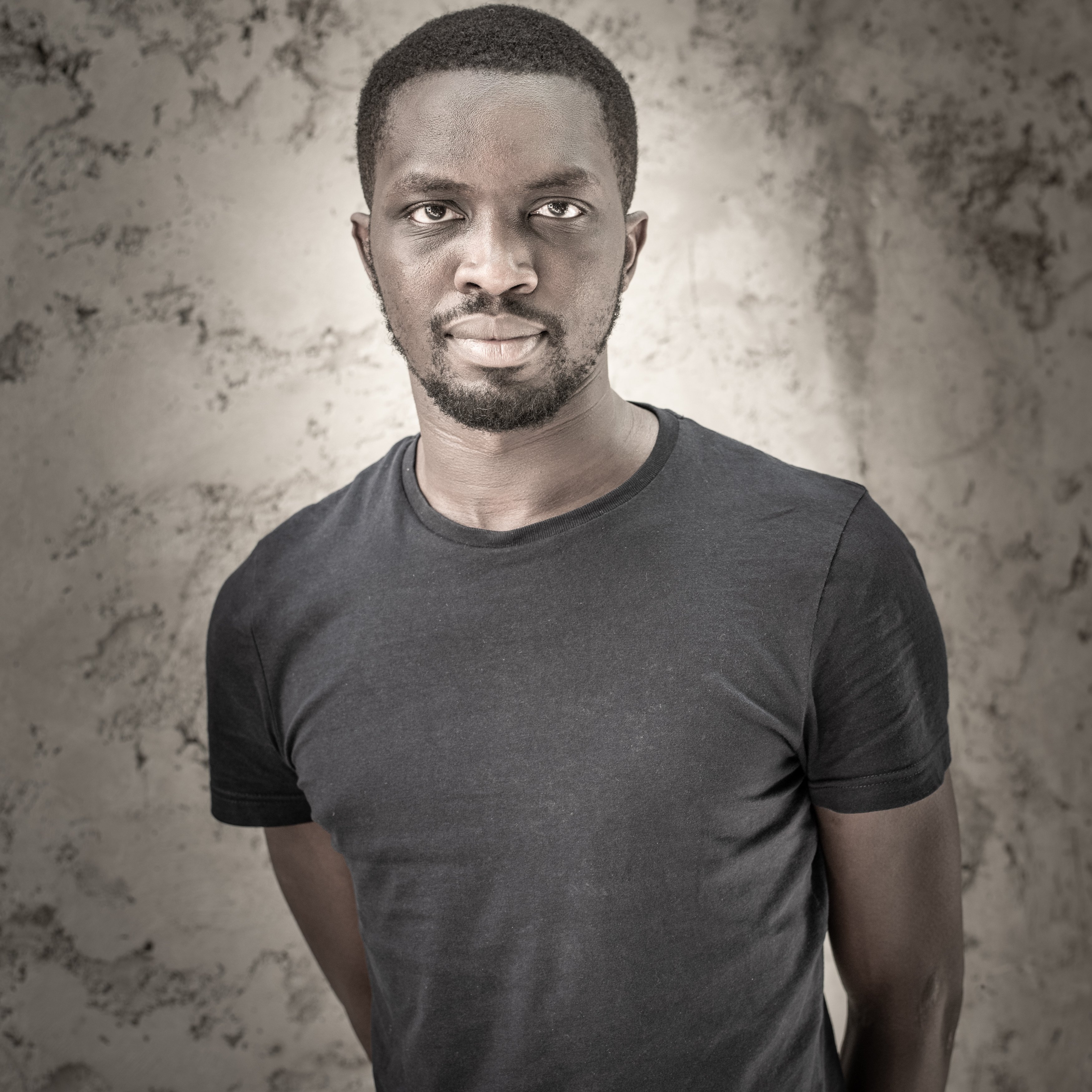 Cette image présente un homme debout, portant un t-shirt noir. Il a la peau foncée et un style de coiffure court. Son expression est sérieuse, et il regarde directement vers l'objectif avec une intensité qui attire l'attention. Le fond de l'image est texturé et neutre, mettant en valeur son visage et son corps. La lumière éclaircit certaines zones, ajoutant de la profondeur et du contraste à l'ensemble.