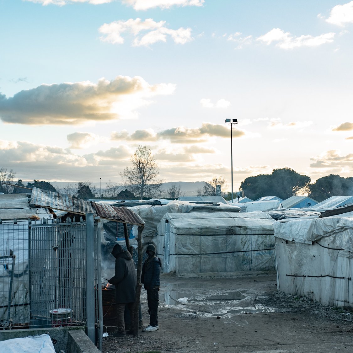 L'image montre un paysage de campement, probablement un bidonville, où des abris temporaires sont installés. Ces structures, couvertes de bâches blanches, sont disposées de manière désordonnée, formant des allées étroites entre elles. Au premier plan, deux personnes se tiennent près d'un feu, suggérant un moment de convivialité ou de chaleur dans un environnement difficile. Le ciel est nuageux, avec des teintes de bleu et d'orange, indiquant une heure de la journée où le soleil se lève ou se couche. En arrière-plan, des arbres et des monts se dessinent, ajoutant une dimension naturelle à ce paysage urbain. L'atmosphère peut sembler mélancolique, mais aussi résiliente, avec des traces de vie et d'activités humaines.