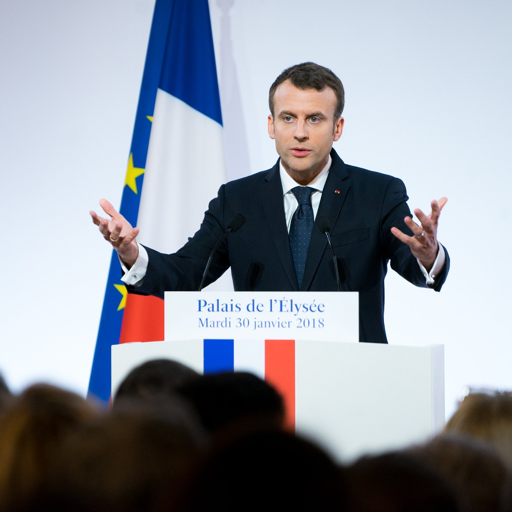 L'image montre un homme debout devant un podium, probablement en train de donner un discours. Il porte un costume sombre avec une cravate, et ses mains sont levées, ce qui indique qu'il met l'accent sur ses propos. En arrière-plan, on aperçoit le drapeau français aux couleurs bleu, blanc et rouge, ainsi qu'une autre bannière européenne. La scène semble se dérouler dans un cadre officiel, peut-être à l'Élysée, avec un public flou et attentif. L'atmosphère est sérieuse et engageante, marqué par le dynamisme du orateur.