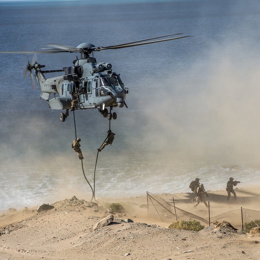 L'image montre un hélicoptère militaire en pleine action, en train de décoller ou de se poser près d'une côte. On peut entendre le bruit des pales qui tournent rapidement, créant un nuage de poussière et de sable autour de lui. À proximité, des soldats descendent en rappel, suspendus à une corde, tandis qu'ils avancent vers une zone de terrain difficile. En arrière-plan, on aperçoit la mer, calme, et une structure qui ressemble à un bâtiment près de la plage. L'atmosphère générale dégage un sentiment d'urgence et d'opération militaire.
