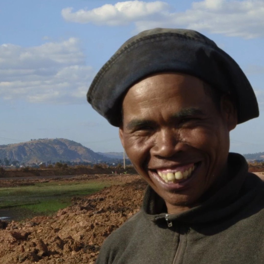 L'image montre un homme souriant se tenant devant un paysage rural. Il porte un chapeau noir et un vêtement sombre. En arrière-plan, on peut voir des collines et un ciel parsemé de nuages blancs. Le sol est terreux, et il y a des zones d'eau, probablement des rizières. L'expression de l'homme dégage de la joie et de la chaleur, ajoutant une touche positive à cette scène naturelle.