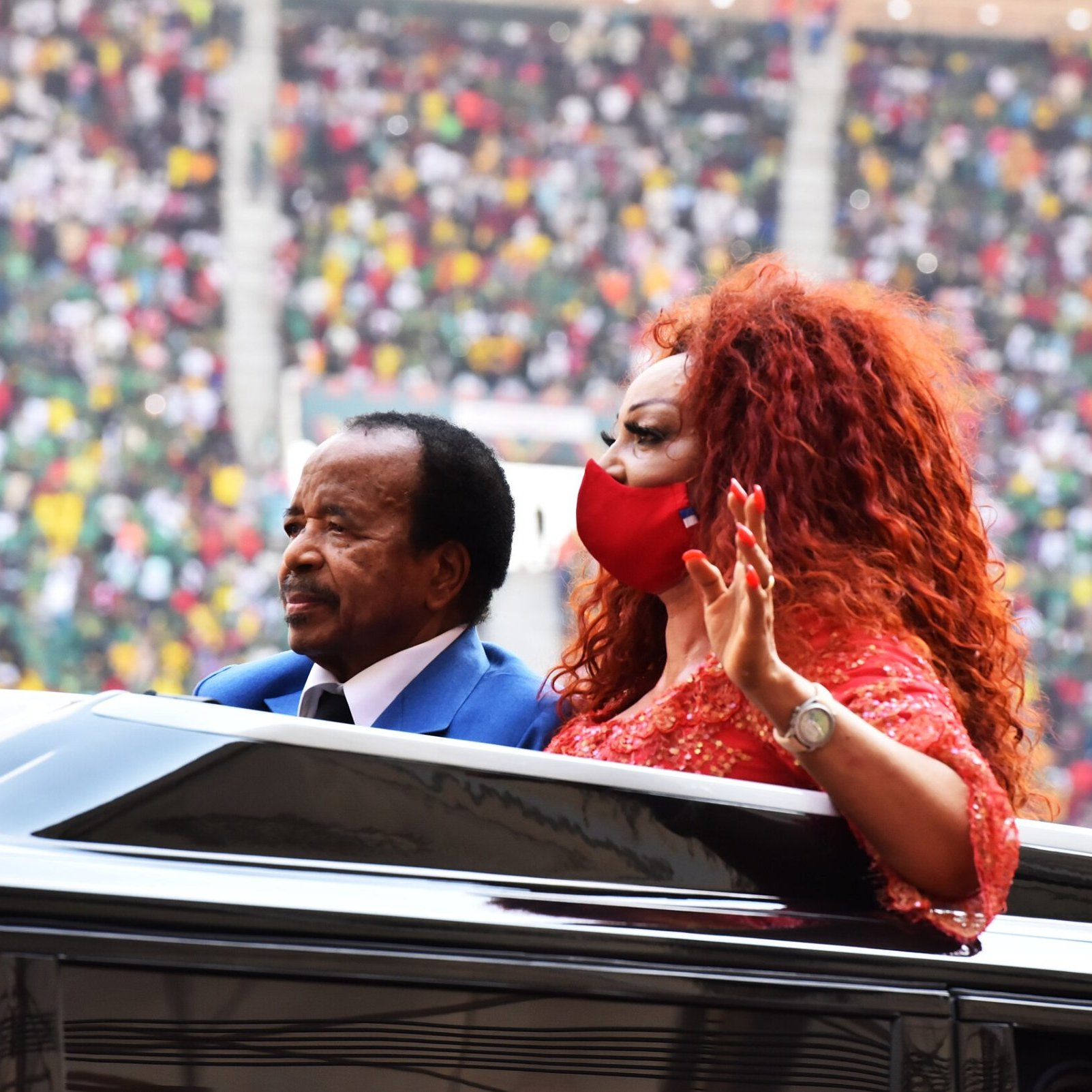 Dans cette image, nous voyons un homme et une femme qui se trouvent à l'intérieur d'une voiture décapotable. L'homme, habillé en costume, a une expression sérieuse et semble saluer la foule. La femme, aux cheveux volumineux et roux, porte une robe brillante et un masque rouge, et elle lève la main en signe de salutation. En arrière-plan, le stade est rempli de spectateurs, avec des couleurs vives de vêtements qui suggèrent un rassemblement festif. L'ambiance générale semble être celle d'un événement important, peut-être une cérémonie ou une célébration.