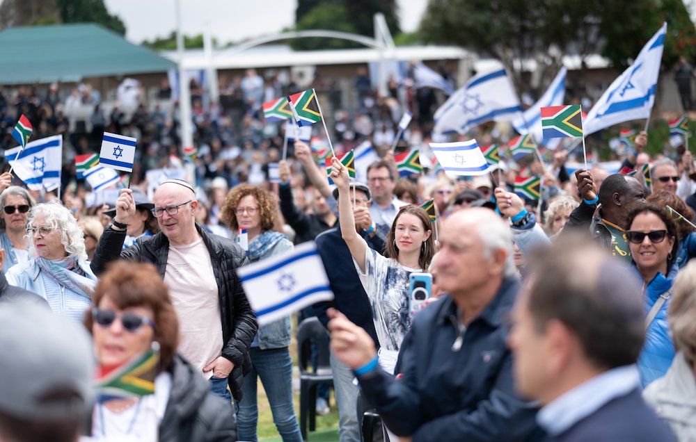 L'image montre une grande foule de personnes rassemblées dans un espace extérieur. Les gens tiennent des drapeaux, principalement des drapeaux israéliens, qui sont blancs et bleus avec l'Étoile de David, ainsi que des drapeaux sud-africains, qui présentent des couleurs vives. L'atmosphère est festive et engagée, avec des visages souriants et des expressions de joie. En arrière-plan, on peut apercevoir des assises et des personnes rassemblées autour d'un événement, suggérant une célébration ou une manifestation significative. Les vêtements des participants sont variés, allant de tenues décontractées à plus habillées, illustrant la diversité de la foule.