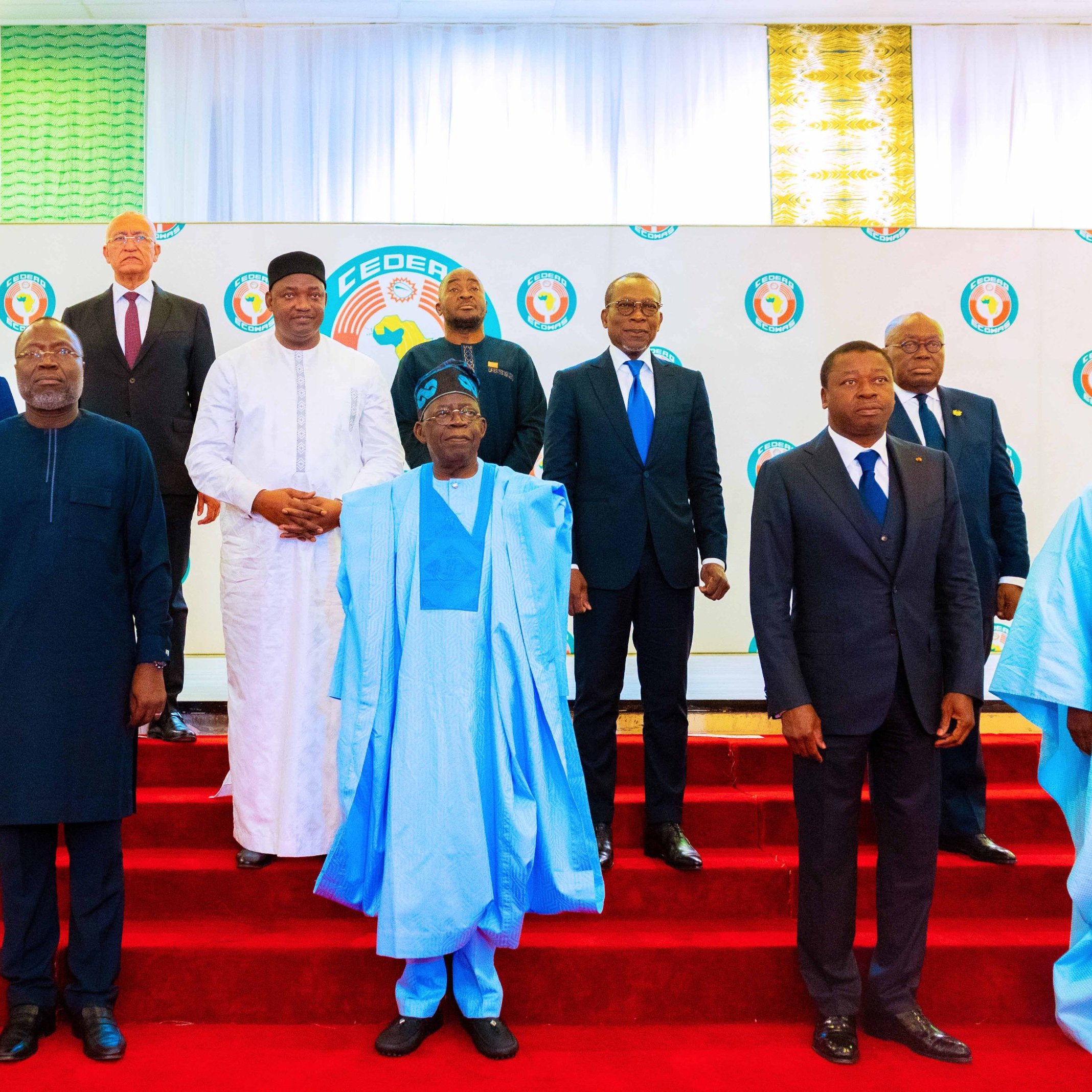 L'image montre un groupe de plusieurs hommes posant ensemble sur une scène. Ils sont vêtus de costumes formels et de tenues traditionnelles. La scène est décorée avec un fond de couleurs vives, comprenant un motif de logo sur la toile blanche. Les hommes se tiennent sur une estrade de couleur rouge, formant une sorte de demi-cercle. Certains portent des vêtements à motif et d'autres des costumes classiques, reflétant une diversité culturelle. L'atmosphère semble officielle, suggérant un événement important ou une réunion.