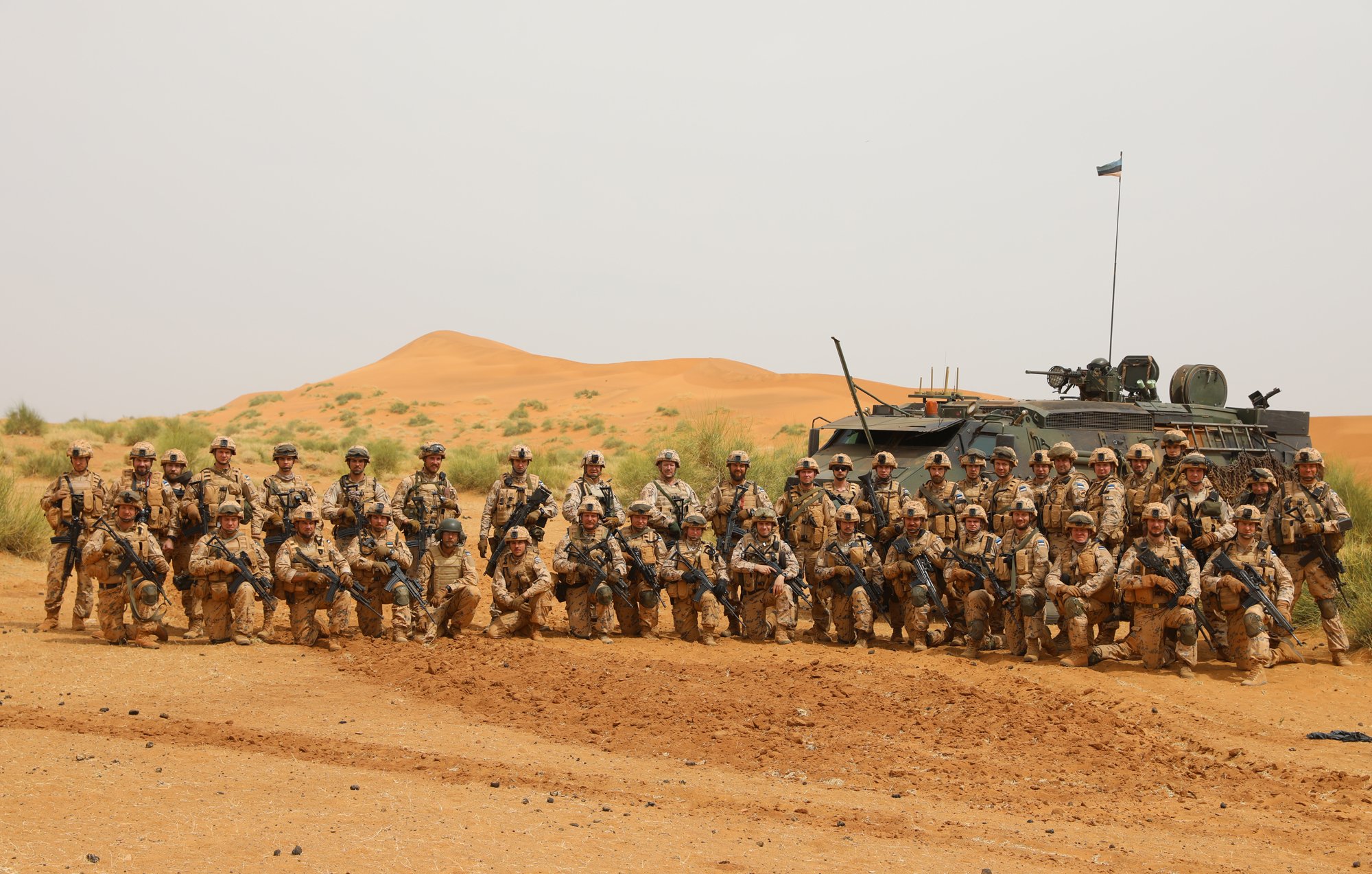 L'image montre un groupe de soldats rassemblés dans un paysage désertique. Au premier plan, on peut voir une trentaine de militaires, tous vêtus de tenues camouflées dans des tons de sable et de marron. Ils se tiennent debout ou accroupis, posant avec des expressions sérieuses. À l'arrière, un véhicule militaire, probablement un blindé, est stationné et porte un drapeau sur son toit. Le fond est marqué par des dunes de sable orangé, typiques des déserts, sous un ciel clair. Cette scène évoque un contexte militaire et une atmosphère de camaraderie entre les soldats.