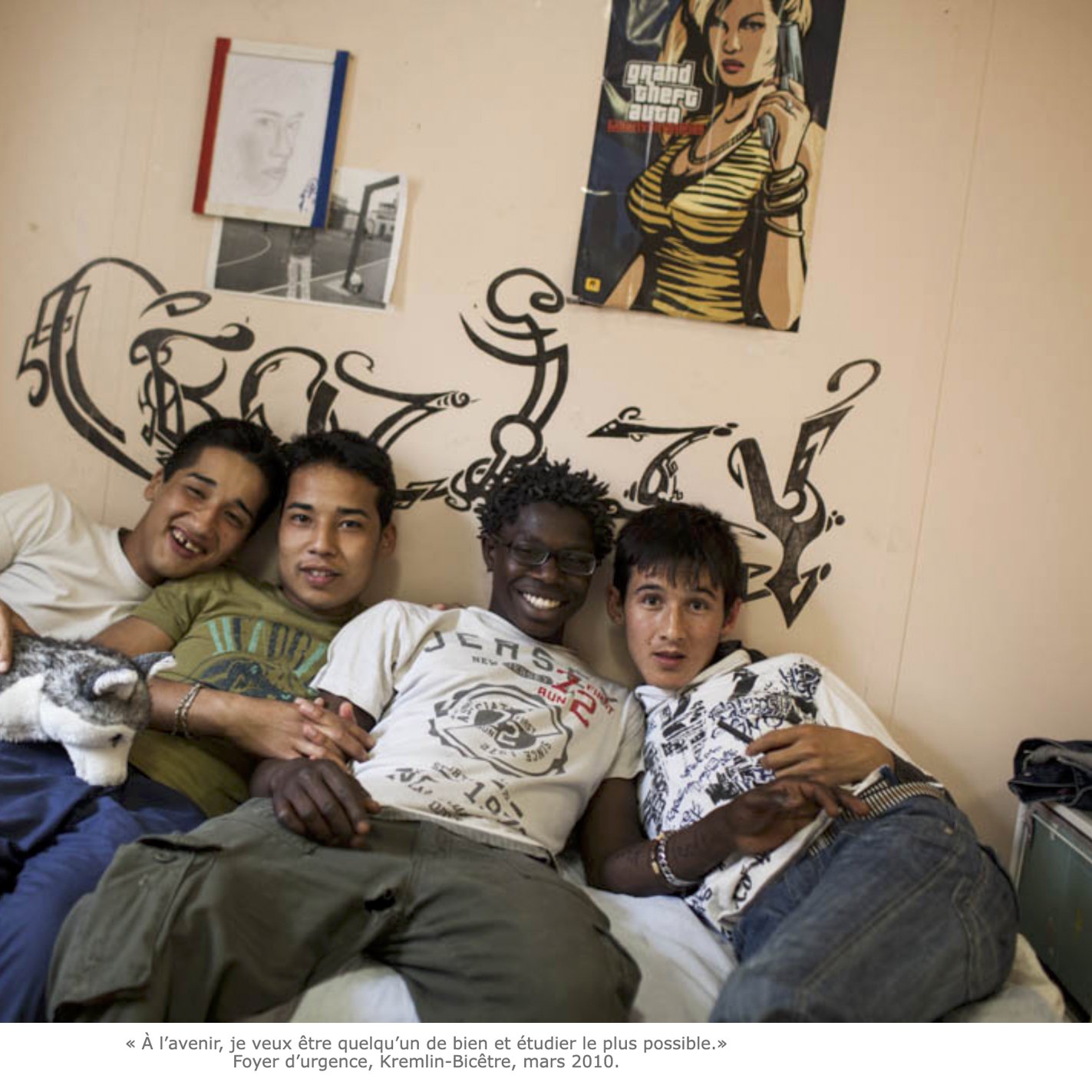 L'image montre quatre jeunes hommes posant ensemble sur un lit dans une chambre. Ils sourient, affichant une ambiance amicale et détendue. La pièce est décorée simplement, avec un mur où l'on peut voir des graffitis. À côté d'eux, un petit chat est blotti. L'un des jeunes tient un ours en peluche. Chaque personne présente une tenue décontractée. Les expressions sur leurs visages suggèrent un moment de complicité et de camaraderie.