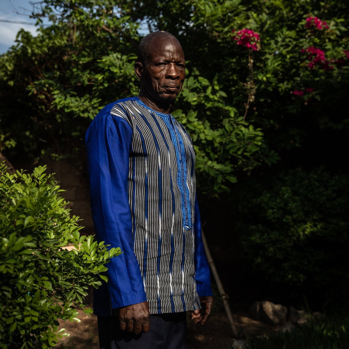 L'image montre un homme debout dans un environnement verdoyant. Il porte une tunique à manches longues, qui est ornée de lignes bleues et de motifs subtils. Son expression est sérieuse et son regard est dirigé vers l'objectif. Autour de lui, on peut apercevoir des plantes luxuriantes, avec des feuilles brillantes et quelques fleurs colorées en arrière-plan. La lumière du soleil filtre à travers les branches, créant une atmosphère tranquille.