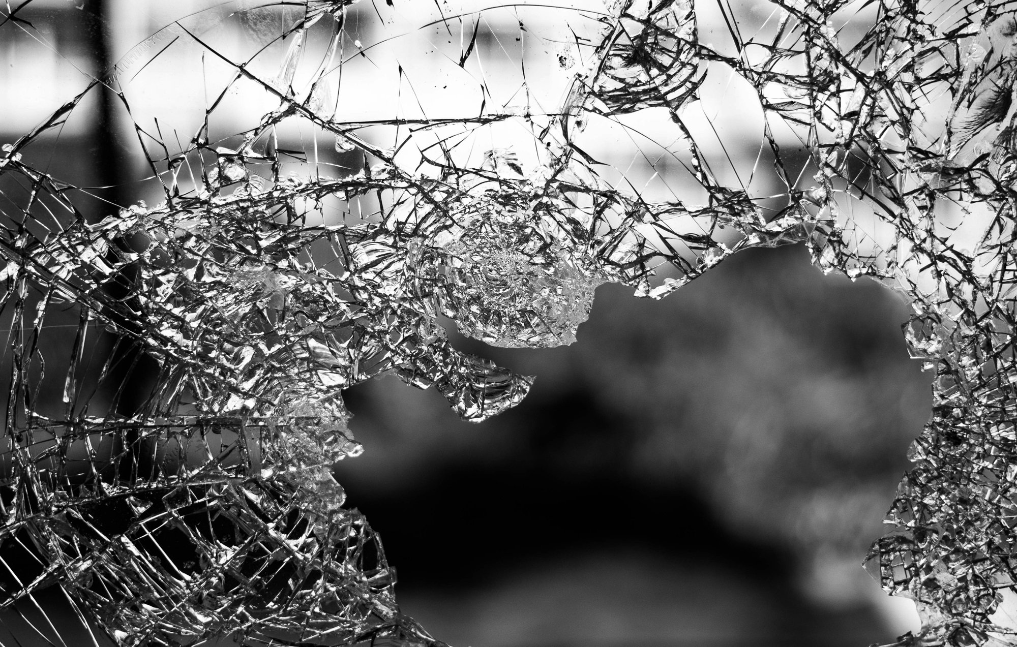 L'image montre un morceau de verre brisé, dont les nombreux éclats forment des motifs irréguliers. On peut ressentir la fragilité et la netteté des bords du verre. La lumière semble traverser certains éclats, créant des jeux d'ombre et de lumière. L'arrière-plan est flou, évoquant une ambiance sombre et mystérieuse. Cette composition souligne la beauté inattendue du verre cassé, tout en évoquant une sensation de vulnérabilité.