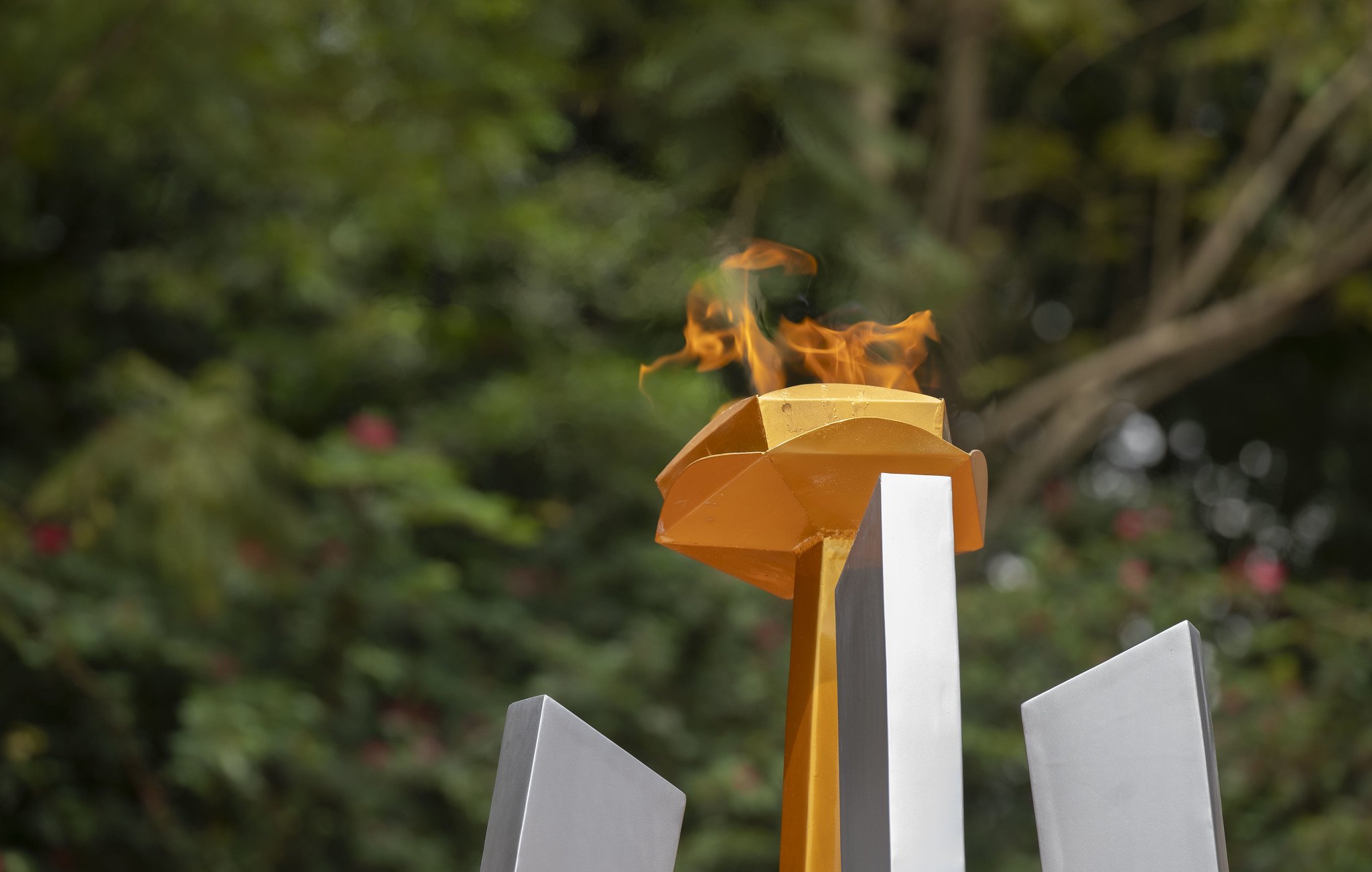 L'image représente une flamme allumée sur un socle jaune, qui surplombe plusieurs structures géométriques grises. La flamme danse avec vivacité, créant une ambiance dynamique, entourée d'un fond verdoyant flou, suggérant la présence d'arbres et de végétation. Cette scène évoque un symbole de passion et de célébration, souvent associé à des événements prestigieux comme les Jeux Olympiques. L'ensemble dégage une impression de force et de détermination.