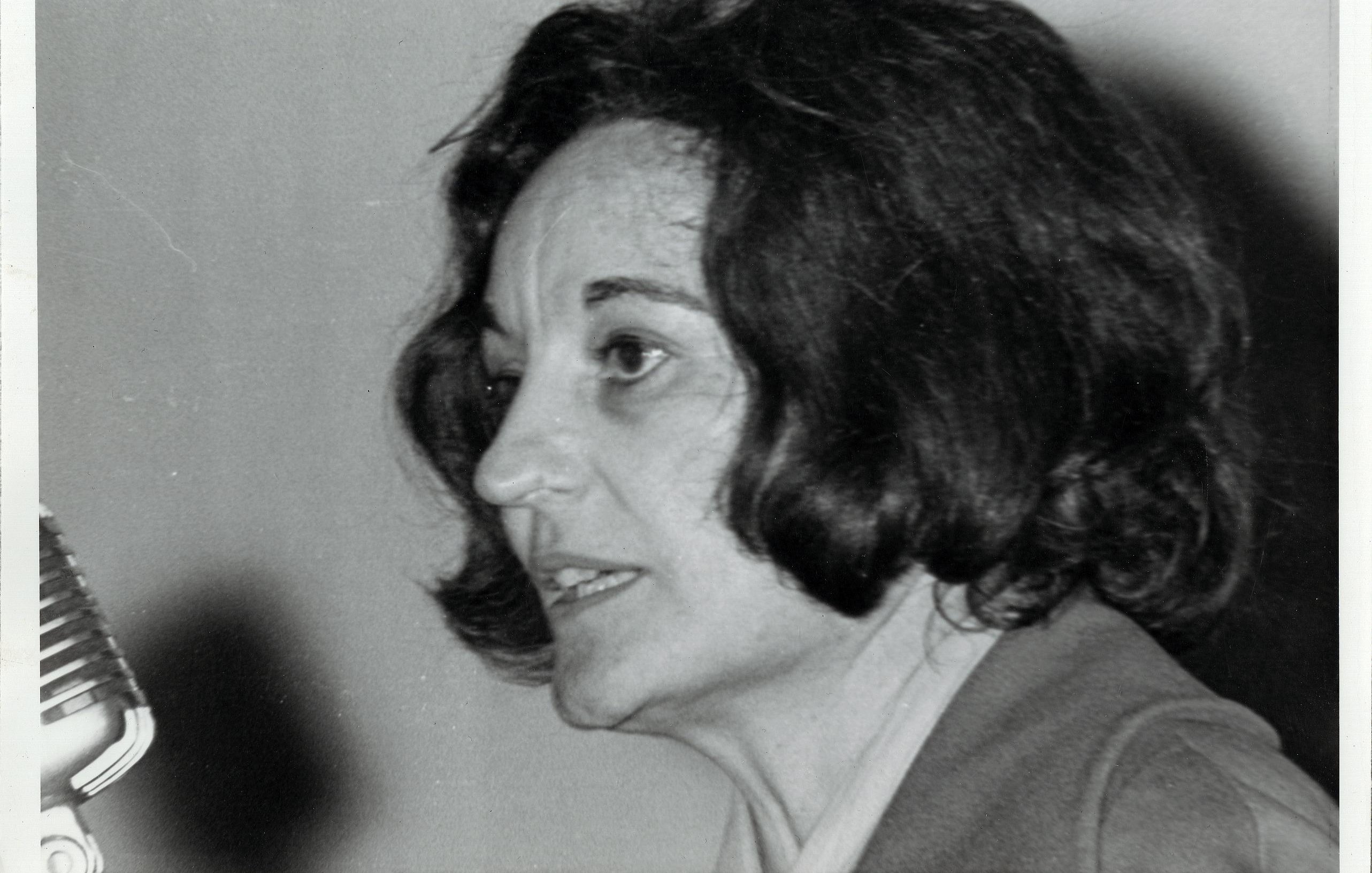 L'image montre une femme avec des cheveux foncés, mi-longs et légèrement ondulés. Elle est en profil, parlant près d'un microphone. Son expression est concentrée, et on peut deviner une certaine passion dans son discours. Elle porte une veste simple, et l'arrière-plan est flou, ce qui met l'accent sur son visage et son expression. L'éclairage est doux, soulignant les traits de son visage.