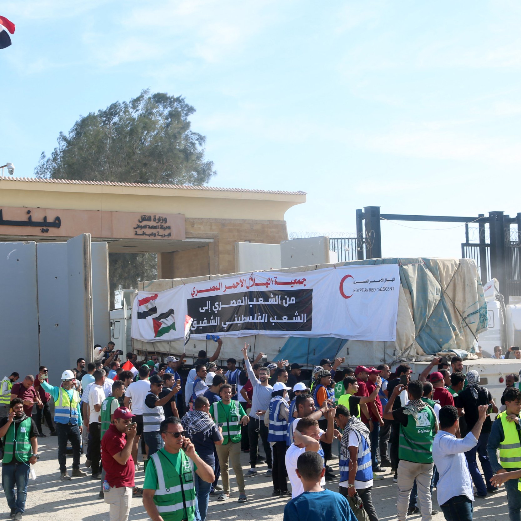 L'image montre une scène animée devant un point de passage, probablement dans une région à fort trafic. On aperçoit des personnes regroupées, certaines portant des gilets de sécurité ou des uniformes, tandis que d'autres tiennent des pancartes. En arrière-plan, on distingue un bâtiment gouvernemental avec des drapeaux en hauteur, ce qui suggère une ambiance officielle ou politique. Les gens semblent engagés dans une sorte de manifestation ou de rassemblement, discutant entre eux et brandissant des affiches qui expriment des revendications. L'atmosphère est festive mais sérieuse, marquée par le dynamisme des participants et les messages affichés.