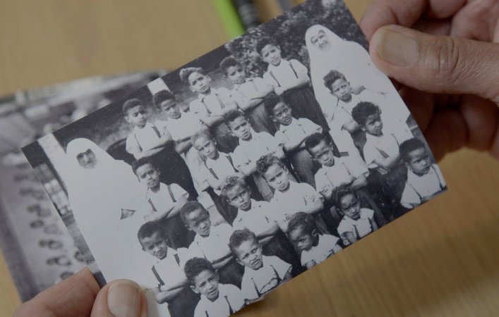 L'image montre une photographie en noir et blanc d'un groupe d'enfants, probablement d'une école ou d'une classe. Les enfants, qui portent des uniformes clairs, sont rassemblés en plusieurs rangées, affichant des expressions diverses. Au centre, une personne vêtue d'une tenue traditionnelle, probablement une enseignante ou une surveillante, se tient parmi eux. L'arrière-plan est flou, mais on peut deviner une ambiance scolaire. Les mains de quelqu'un tiennent la photo avec précaution, indiquant son importance personnelle ou sentimentale.