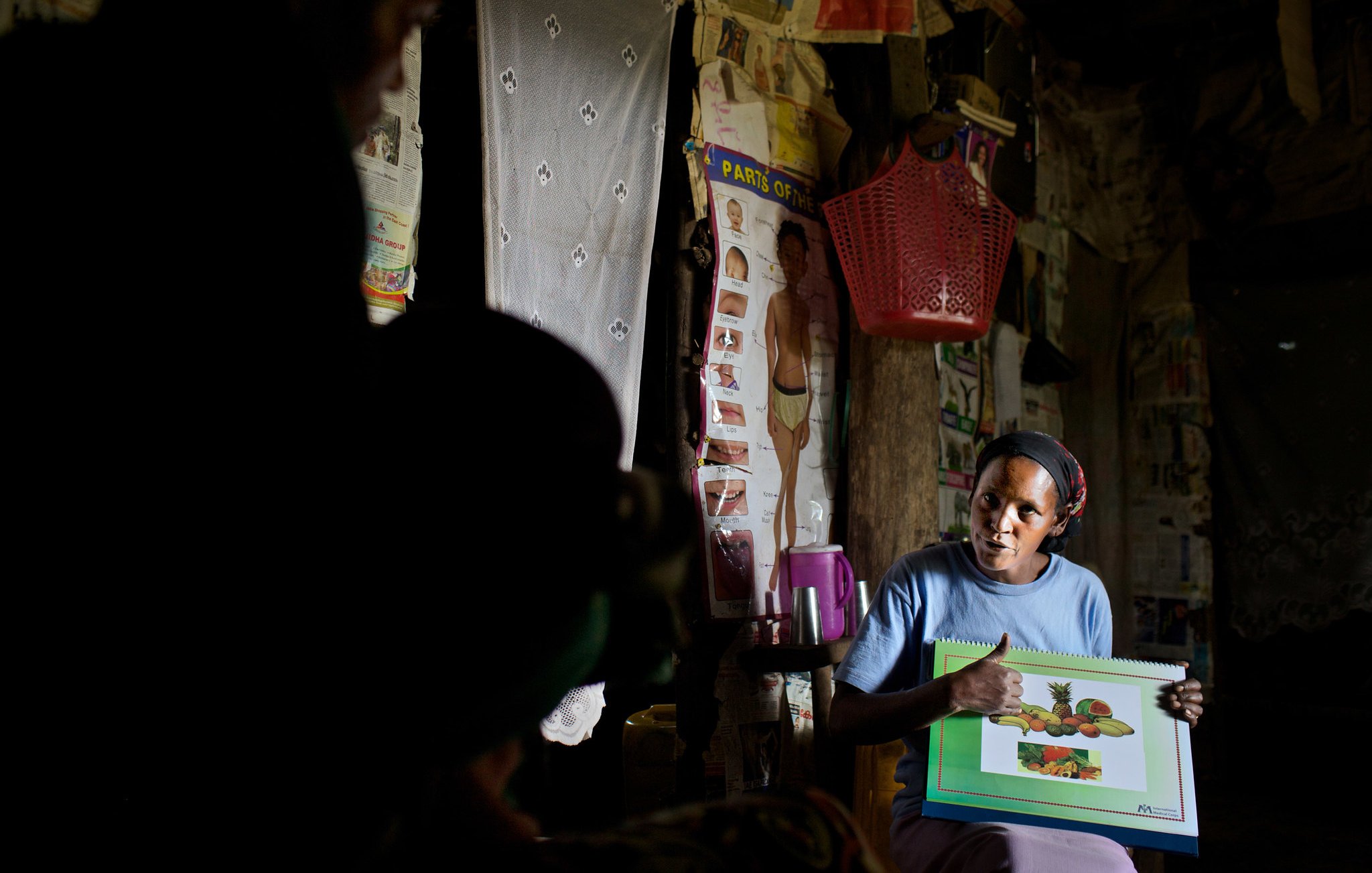 L'image montre une femme assise dans une pièce sombre, entourée de murs décorés d'affiches et de tissus. Elle tient un grand carnet avec des illustrations de fruits, qu'elle montre avec enthousiasme. Sa tenue est simple, et elle a l'air concentrée sur son explication. À l'arrière-plan, une silhouette d'une autre personne est visible, ajoutant à l'atmosphère d'échange. La scène évoque un moment d'apprentissage ou de partage d'informations sur les fruits.