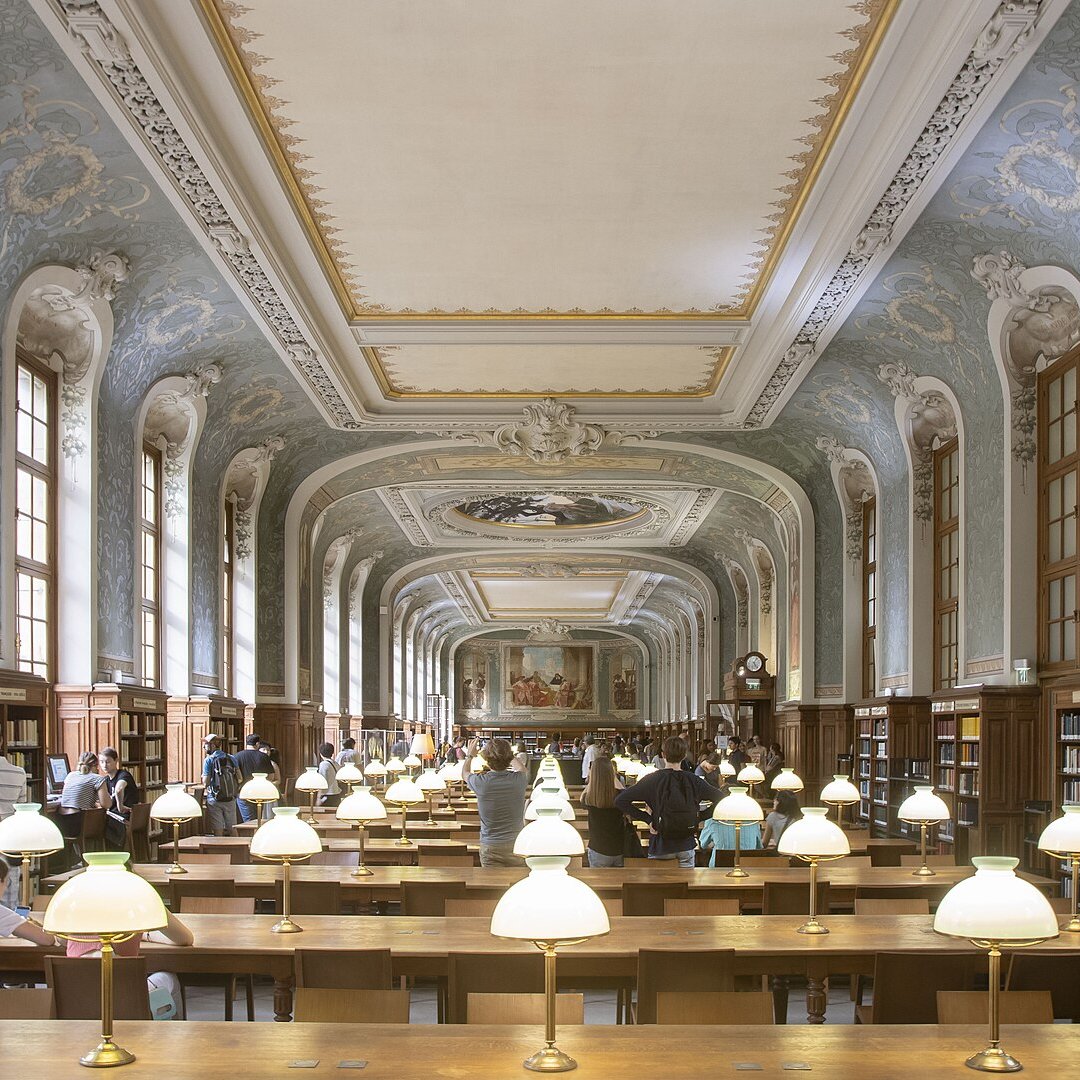 Imaginez une vaste bibliothèque, où l'air est chargé de silence et de connaissance. L'espace est long et majestueux, avec un plafond haut orné de décorations raffinées et de motifs artistiques. Les murs sont bordés de grandes fenêtres, permettant à la lumière naturelle d'inonder la pièce. Des lampes de table éclatent par leur lumière douce sur de longues tables en bois, disposées en rangées. Ces tables accueillent des visiteurs plongés dans leurs livres et leurs pensées. Au fond, un grand mur est décoré de fresques, ajoutant une note d'élégance à l'ensemble. Les étagères en bois, pleines de livres soigneusement rangés, s'élèvent majestueusement sur les côtés, évoquant un immense trésor de savoir à portée de main. Les gens se déplacent doucement, parlant à voix basse, ajoutant une ambiance de respect et de tranquillité. Cette bibliothèque semble être un sanctuaire pour les amateurs de lecture et de culture.