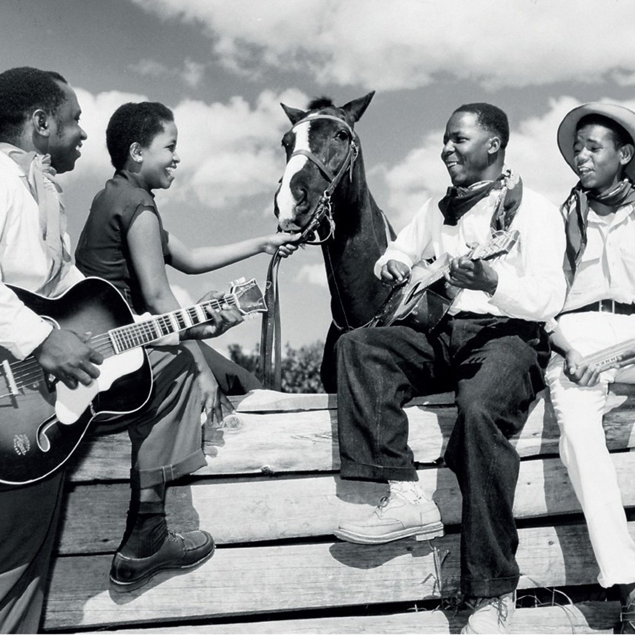 L'image représente un groupe de quatre personnes en noir et blanc, assises autour d'une barrière en bois, sous un ciel partiellement nuageux. Deux hommes et deux femmes sont présents. L'un des hommes joue de la guitare, tandis que l'autre est assis sur un tronc, tenant un instrument à cordes, probablement un banjo ou un ukulélé. Les deux femmes semblent interagir avec l'un des hommes et un cheval qui est également présent, la tête tournée vers eux. Elles affichent des expressions joyeuses, créant une atmosphère conviviale et détendue. Les vêtements des personnages varient : l'un porte un chapeau de cowboy et les autres portent des vêtements simples, typiques de l'époque. L'image évoque un moment de camaraderie et de musique, suggérant un lien fort entre les personnes et la nature environnante.