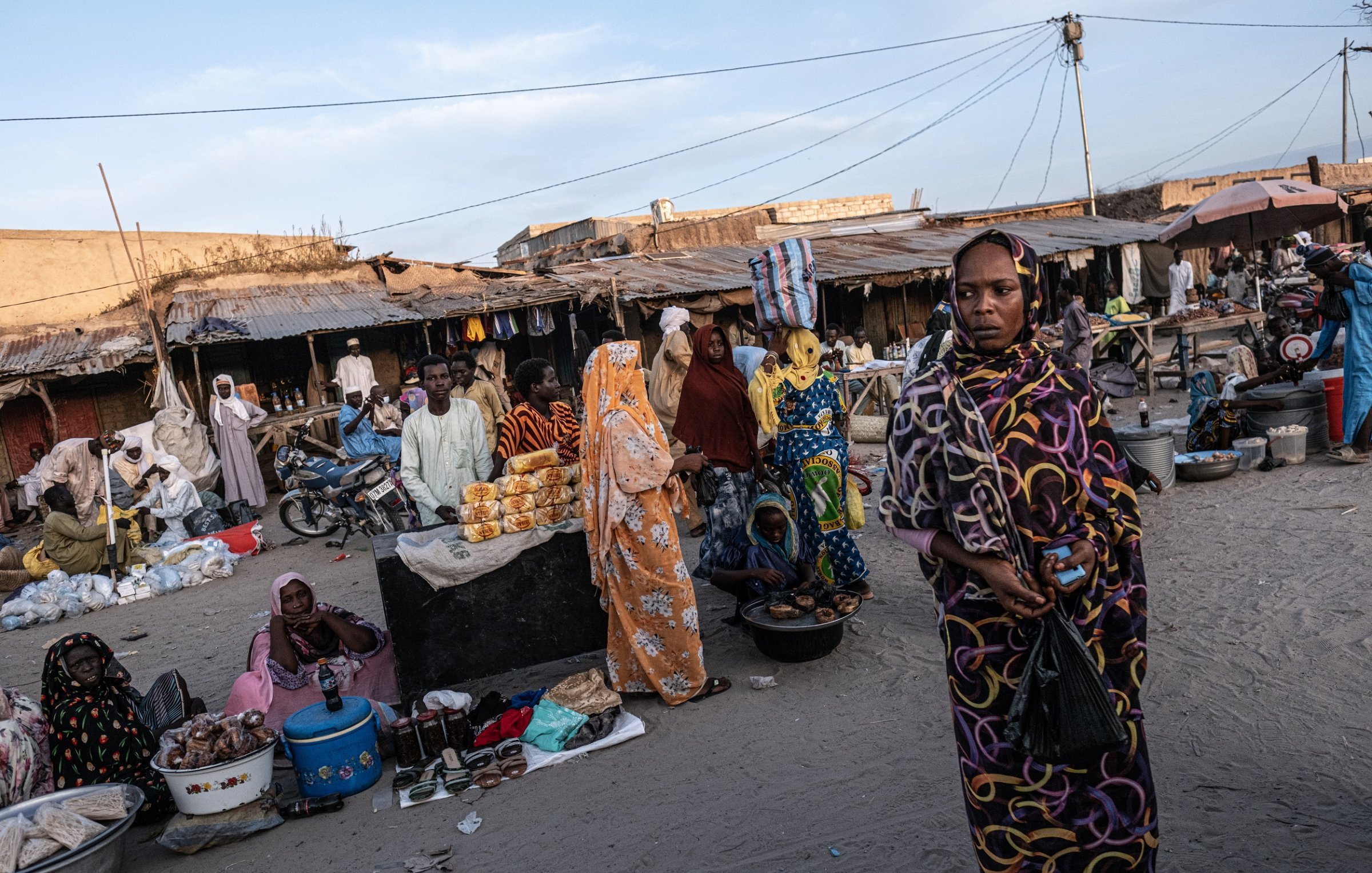 Dans cette scène animée d'un marché, on peut ressentir une atmosphère vibrante. Le sol est recouvert de sable, et plusieurs stands sont disposés tout autour. Des gens sont rassemblés, certains vêtus de vêtements colorés, offrant divers produits alimentaires, tels que des fruits et des pâtisseries. Les femmes, en particulier, attirent l'attention avec leurs robes chatoyantes et leurs voiles. On peut aussi entendre des murmures de conversations, des rires et le bruit de petits véhicules, comme des motos, qui se déplacent à travers le marché. L'air est imprégné d'odeurs variées de nourriture épicée et de douceur, créant une expérience sensorielle unique.