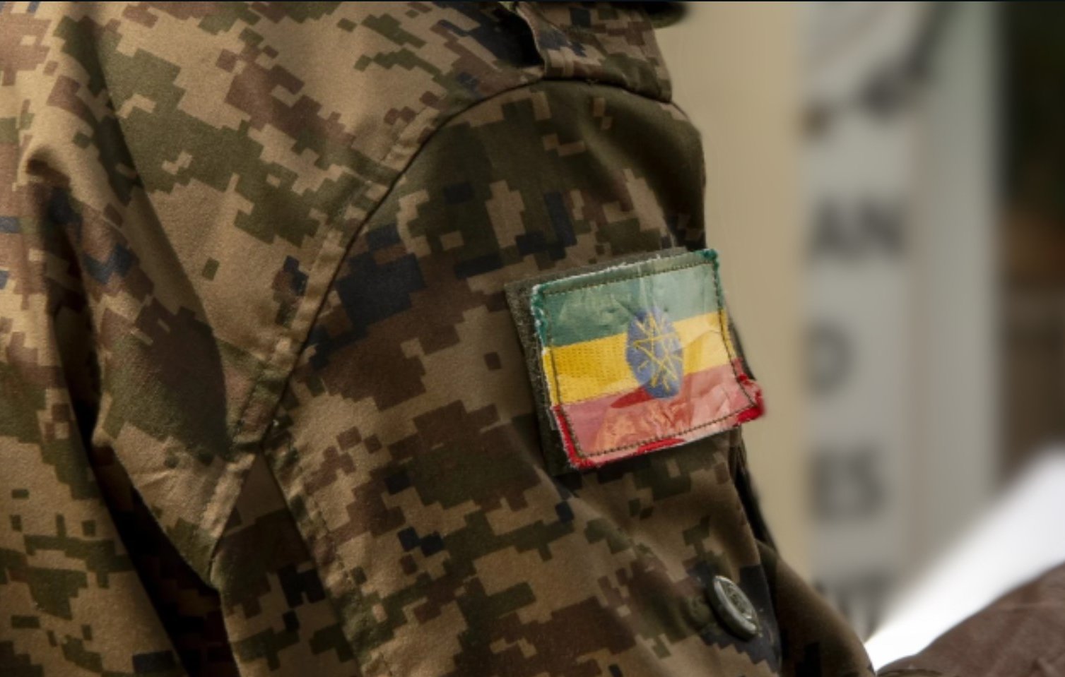 The image features a close-up of a military uniform with a camouflage pattern. Prominently displayed on the arm is a patch showing the flag of Ethiopia, which consists of horizontal stripes in green, yellow, and red, with a blue circle and a yellow star in the center. The fabric of the patch appears worn, adding to its character.