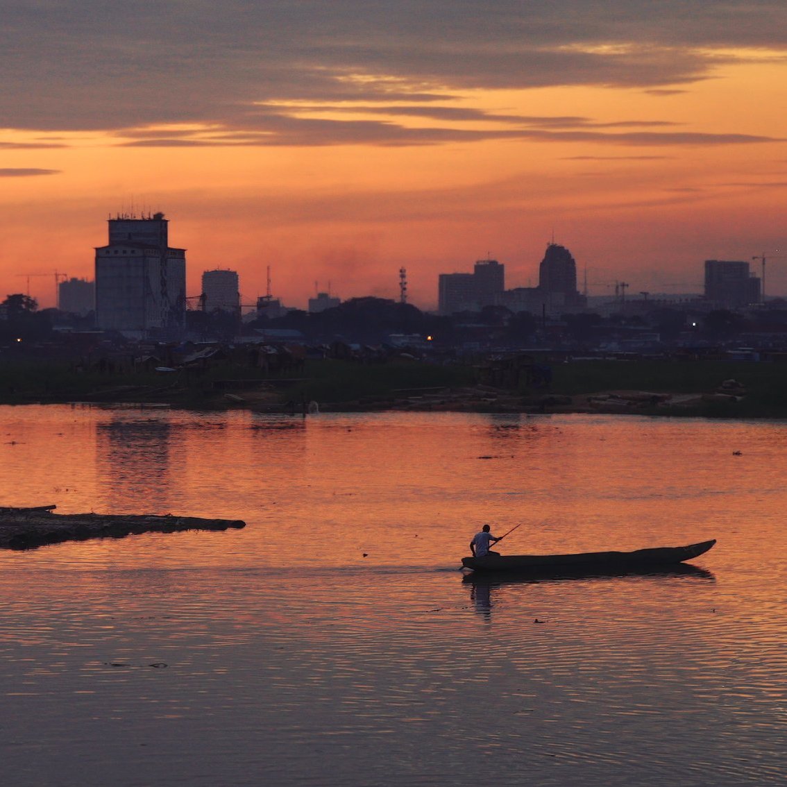 L'image dépeint un paysage paisible au crépuscule. Le ciel est teinté de nuances chaudes d'orange, de rose et de violet, créant une atmosphère calme et sereine. À l'avant, un homme marche au bord de l'eau, tandis qu'un autre navigue tranquillement sur un petit bateau. La surface de la rivière reflète les couleurs du ciel, ajoutant à la beauté de la scène. En arrière-plan, on aperçoit des silhouettes de bâtiments urbains, suggérant la proximité d'une ville. L'ensemble évoque une sensation de tranquillité et de connexion avec la nature, tout en étant en harmonie avec l'activité humaine.