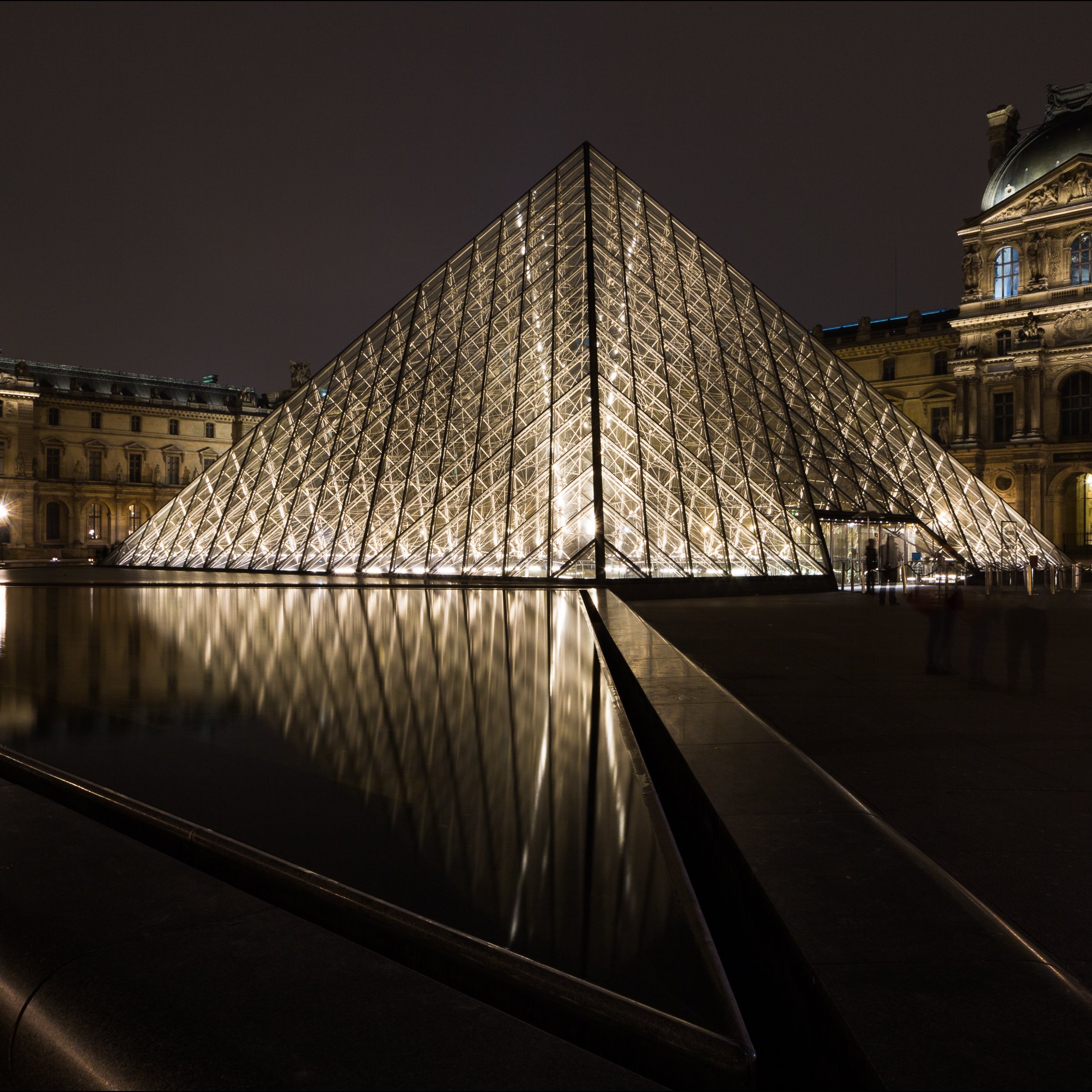 L'image montre le célèbre musée du Louvre à Paris, avec sa pyramide en verre illuminée au centre de la scène. La pyramide, qui est moderne et de forme géométrique, se dresse majestueusement au-dessus d'un bassin d'eau calme qui en reflète la lumière. À gauche, on aperçoit l'architecture historique du musée, avec ses pierres anciennes et des détails raffinés. La scène est plongée dans une ambiance nocturne, où les lumières chatoyantes se mêlent à l'obscurité, créant un contraste saisissant entre le moderne et le classique. Le reflet dans l'eau ajoute une dimension poétique à l'ensemble.