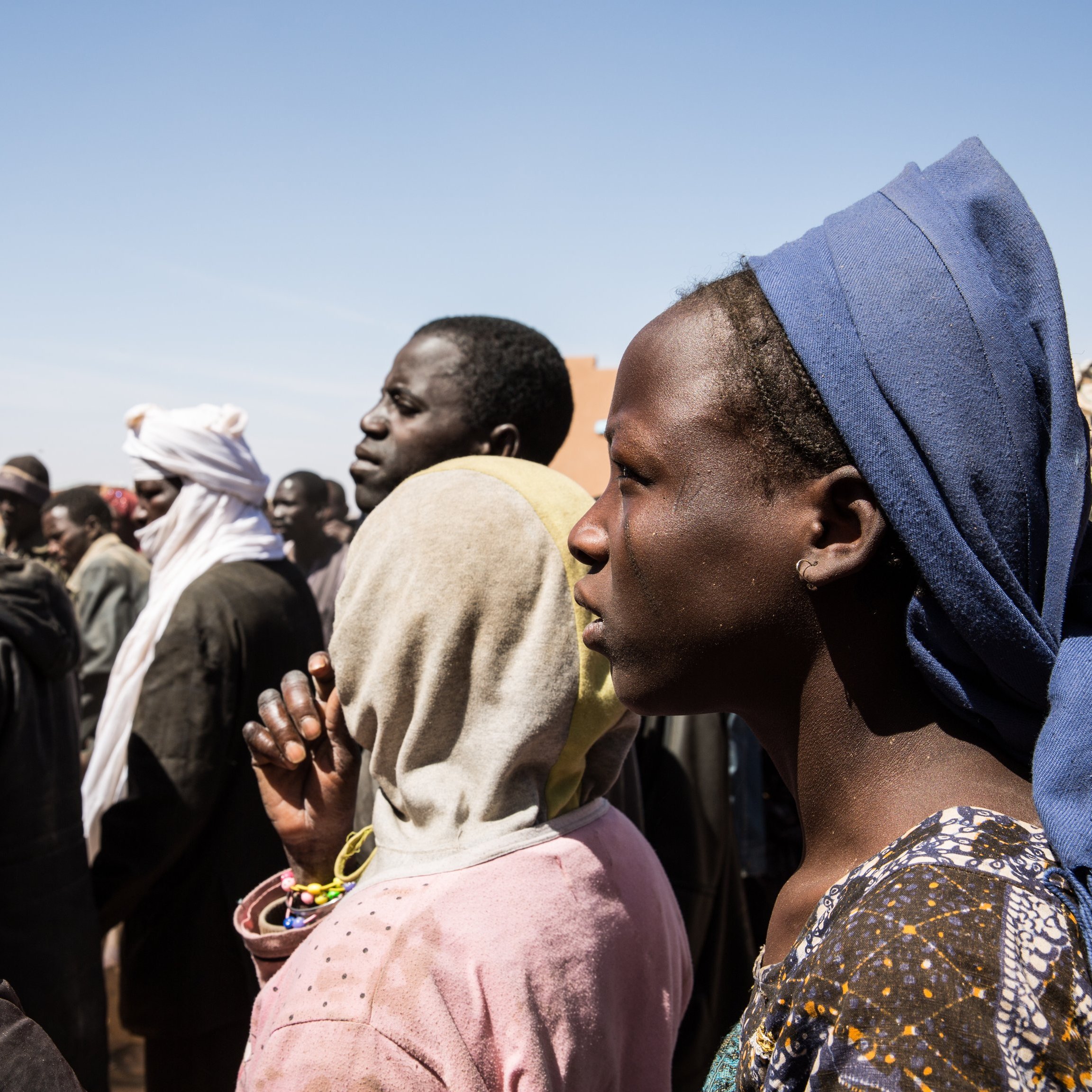 L'image montre un groupe de personnes se tenant debout, la plupart tournées vers un même point. On peut distinguer des hommes et des femmes de différents âges, habillés de vêtements variés, souvent colorés et traditionnels. Certaines personnes portent des couvertures sur la tête ou des scarves. L'expression sur leurs visages semble sérieuse, indiquant peut-être une attente ou une attention particulière. L'arrière-plan présente un ciel clair, suggérant une journée ensoleillée, et des structures émergent faiblement en arrière-plan, ajoutant à l'environnement d'un rassemblement. Le moment semble chargé d'émotions et de tension collective.