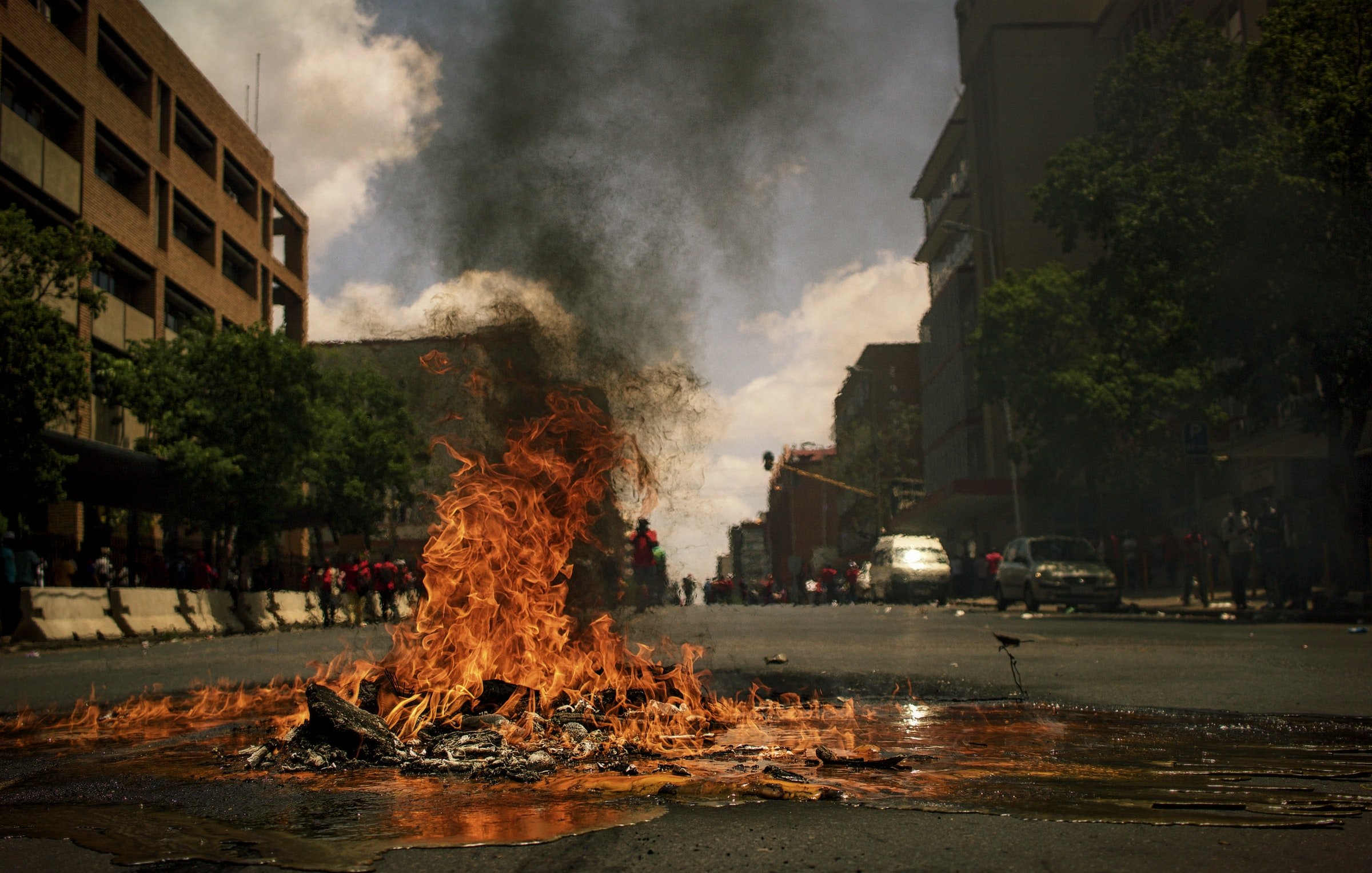 L'image montre une rue animée où un feu brûle au centre. Des flammes vives et tourbillonnantes s'élèvent de ce qui semble être des débris sur le sol. Une épaisse fumée noire s'échappe vers le ciel, obscurcissant partiellement la lumière du jour. On peut apercevoir des bâtiments sur les côtés de la rue, ainsi qu'un groupe de personnes au fond, créant une atmosphère tendue et dynamique. L'air est probablement chargé d'odeurs de brûlé, et le son du crépitement du feu se mêle à une ambiance vibrante de manifestation ou d'agitation.
