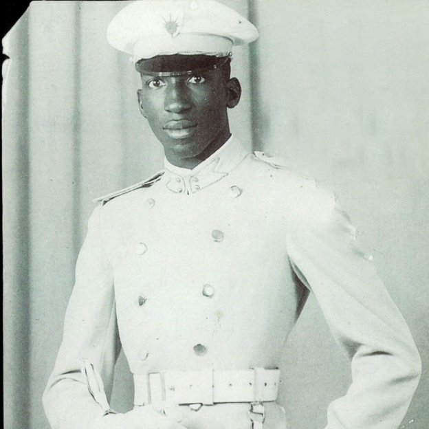 L'image montre un jeune homme en tenue militaire. Il porte un uniforme blanc qui se compose d'une chemise et d'un pantalon assortis, avec une ceinture qui souligne sa taille. Sur sa tête, il a une casquette blanche ornée d'un insigne. Son expression est sérieuse et déterminée. L'arrière-plan est neutre, probablement un drap ou un fond uni, ce qui met en valeur sa silhouette et son uniforme.