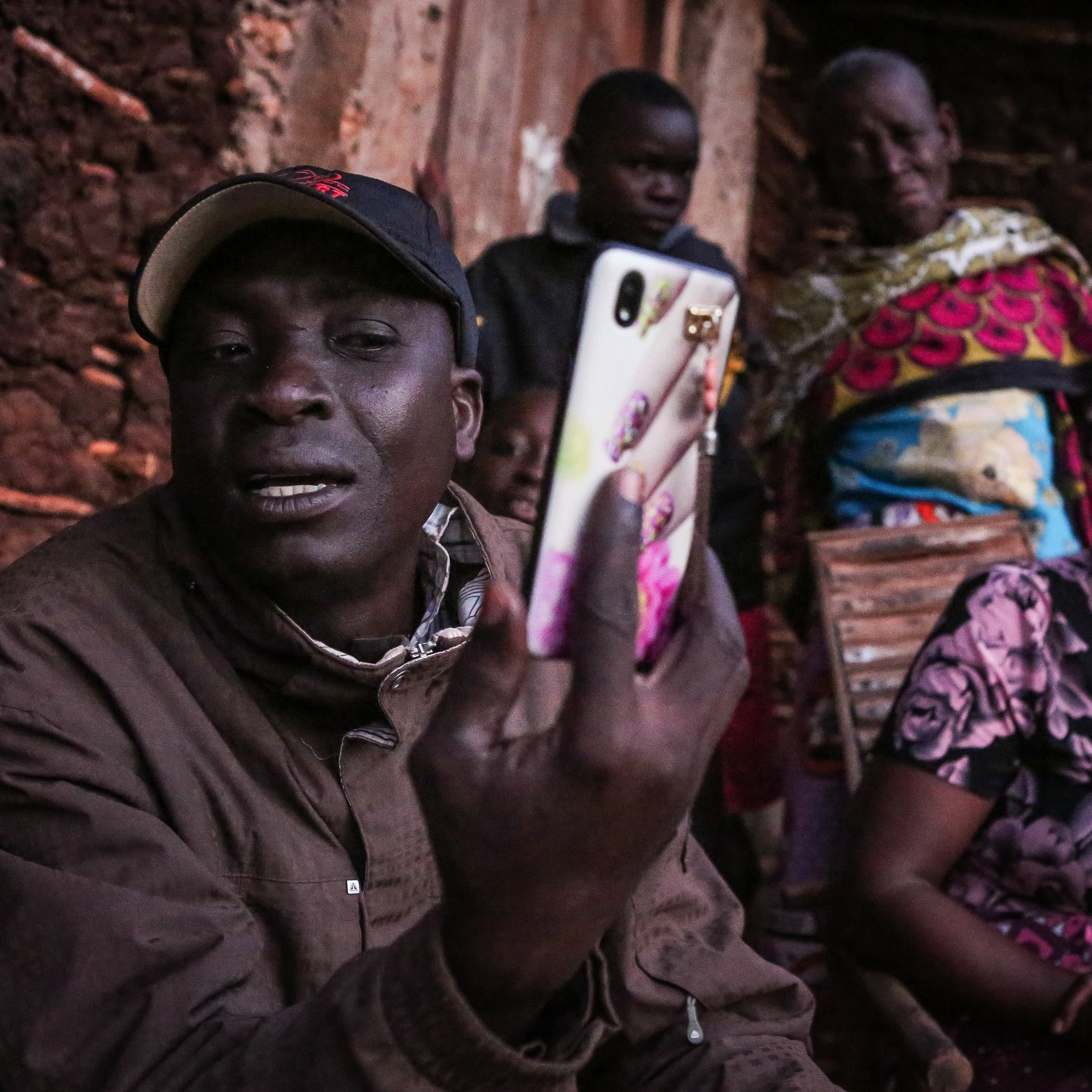 Cette image présente un groupe de personnes rassemblées à l'extérieur d'une maison. Au premier plan, un homme tient un téléphone portable et semble communiquer ou montrer quelque chose à ses interlocuteurs. Il porte une casquette et un manteau. À l'arrière-plan, plusieurs femmes regardent avec attention. Elles sont vêtues de vêtements colorés et affichent des expressions d'intérêt ou de curiosité. L'environnement est simple, avec des murs en terre crue et un éclairage qui suggère qu'il pourrait être tard dans la journée. L'atmosphère semble conviviale et axée sur la communication.