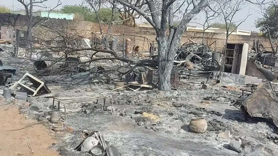 L'image montre un paysage de destruction et de désolation. Au premier plan, le sol est noirci par des cendres et des débris, témoignant d'un récent incendie. Des branches d'arbres calcinées s'élèvent vers le ciel, tandis que des objets métalliques, probablement des meubles ou des ustensiles, sont dispersés autour. On peut voir des pots brisés et des restes de ce qui semblait être une habitation ou un lieu de vie. L'atmosphère est sombre, reflétant la perte et la désolation causées par cet incident.