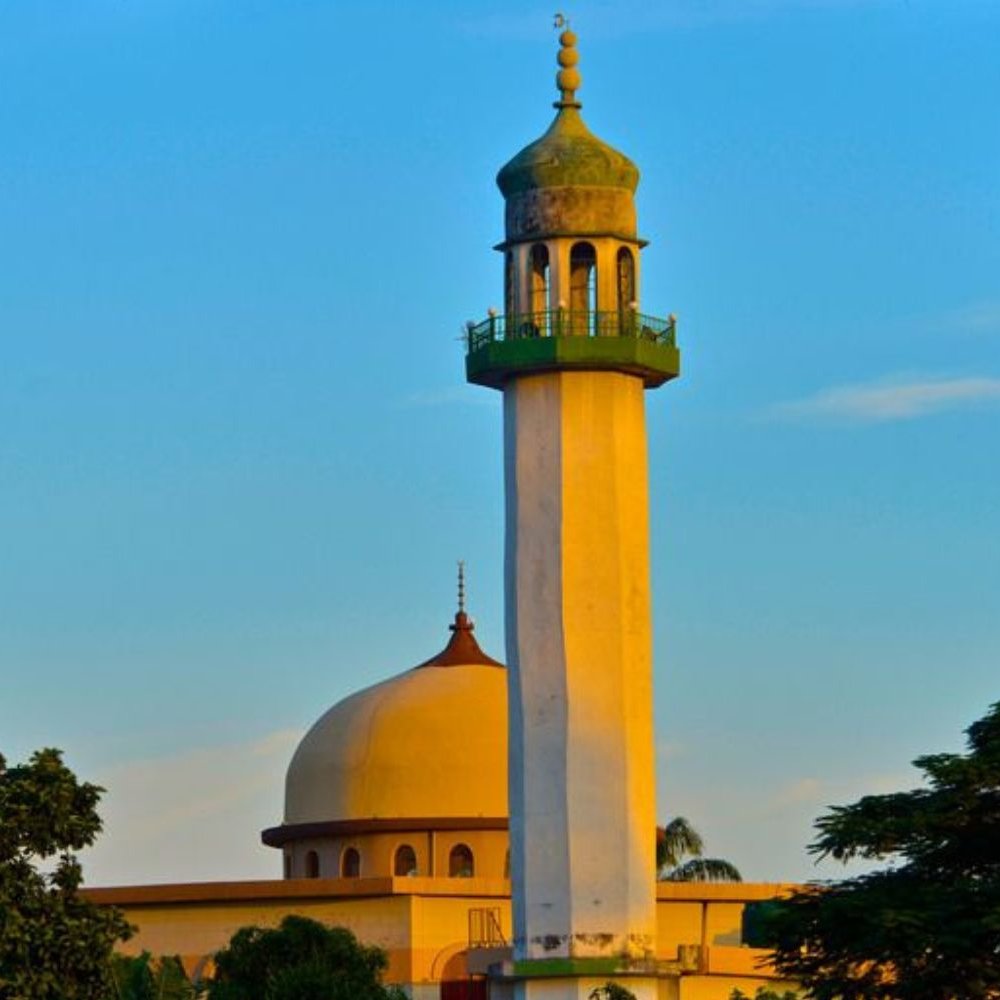 L'image montre un groupe de personnes en train de prier ensemble. Au centre, un homme en costume bleu se tient les mains jointes, la tête légèrement inclinée, montrant une attitude de recueillement. Autour de lui, plusieurs hommes portent des vêtements traditionnels, incluant des turbans et des chapeaux décorés. Tous semblent concentrés et tournés vers l'avant, les yeux fermés ou la tête baissée, dans un moment de méditation ou de prière. L'ambiance est empreinte de sérénité et de respect.