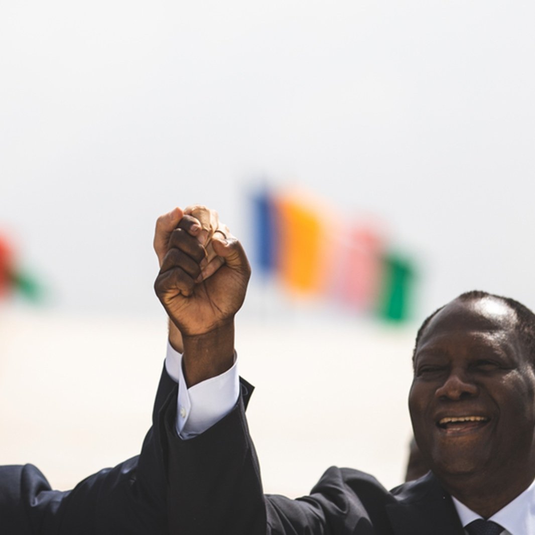 Imaginez une scène marquée par un moment de camaraderie et d'unité. Deux hommes se tiennent côte à côte, levant la main droite en l'air, leurs poings serrés, symbolisant la force et la solidarité. À gauche, on perçoit un homme avec une coupe de cheveux soignée, vêtu d'un costume sombre, tandis que l'homme à droite, également en costume, affiche un grand sourire chaleureux. En arrière-plan, plusieurs drapeaux flottent doucement au vent, représentant différentes nations. L'atmosphère est festive et pleine d'espoir.