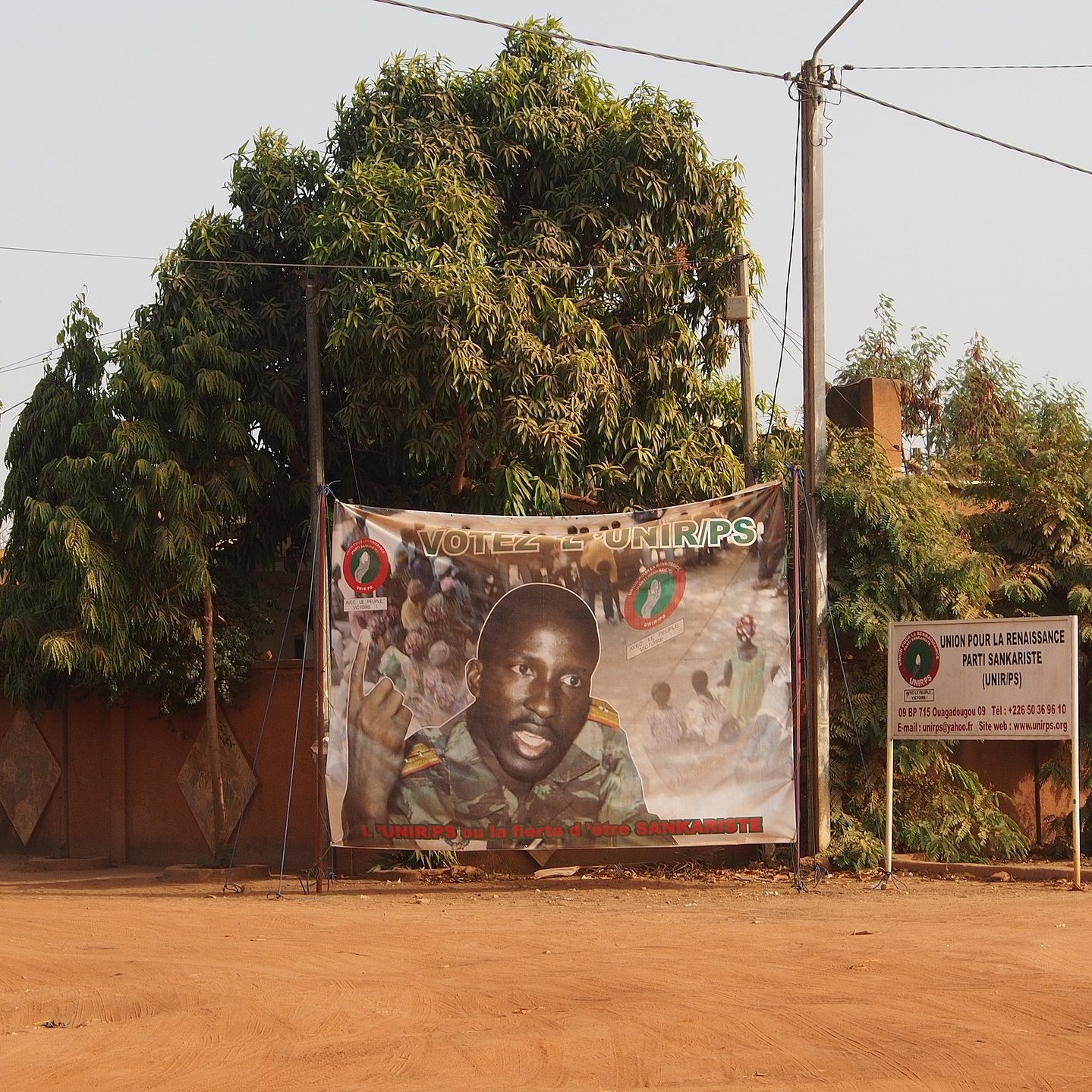 L'image présente une rue déserte d'un quartier, où le sol est recouvert de terre ocre. À gauche, un grand arbre luxuriant, avec des feuilles vertes, apporte une touche de nature. Au centre, une affiche enroulée sur un grillage présente le portrait d'un homme, probablement un candidat politique, souriant et regardant vers l'objectif. L'affiche est ornée de divers motifs colorés et de slogans. À droite, on peut distinguer un panneau d'information sur les élections. Les murs en arrière-plan sont peints dans des tons chauds, contribuant à l'atmosphère locale et authentique de l'endroit.